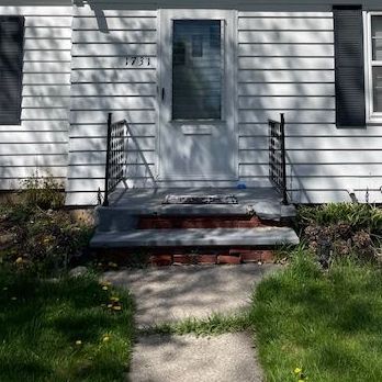 Front entrance of a house with white siding, concrete steps, black railings, and a walkway bordered by grass.