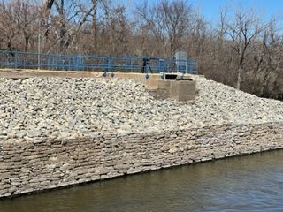 Stone retaining wall along a riverbank with blue railings and leafless trees in the background