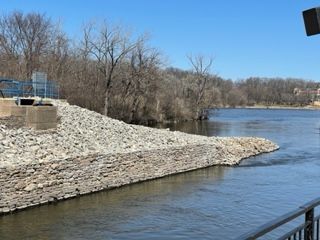 Rock-lined riverbank with a concrete dam edge beside calm blue water and leafless trees