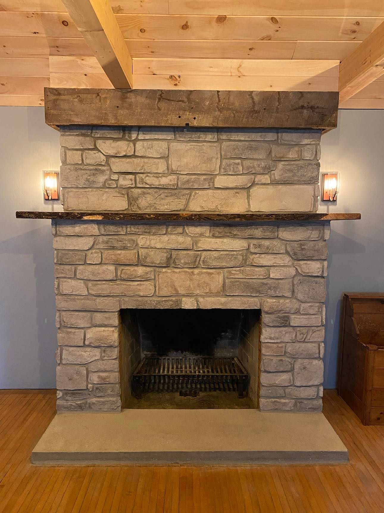 Stone fireplace with wooden mantel in a room, lit wall sconces, and exposed wood ceiling beams