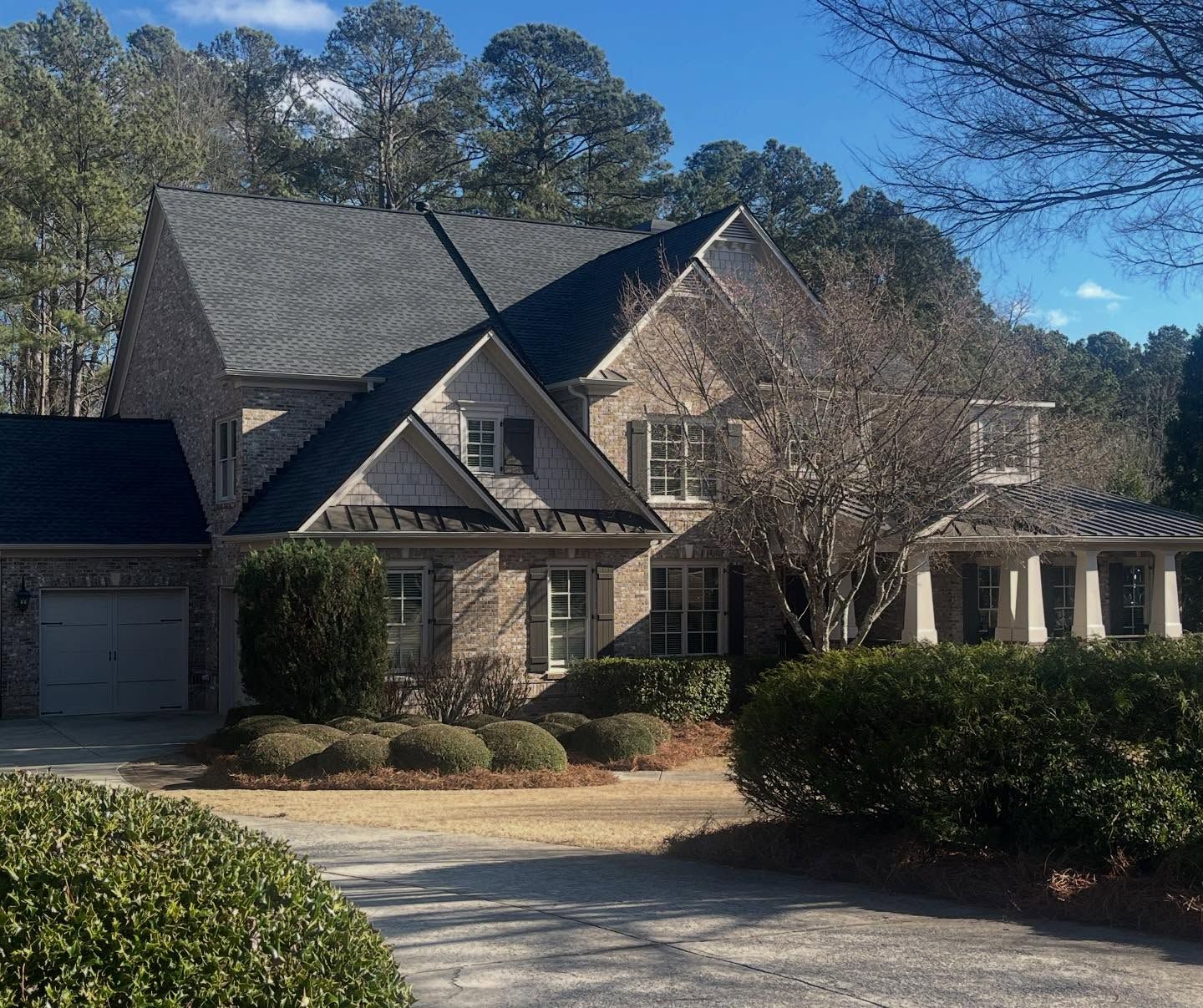 A two-story brick house with a dark gabled roof, tall pine trees in the background, and a driveway with landscaped bushes.