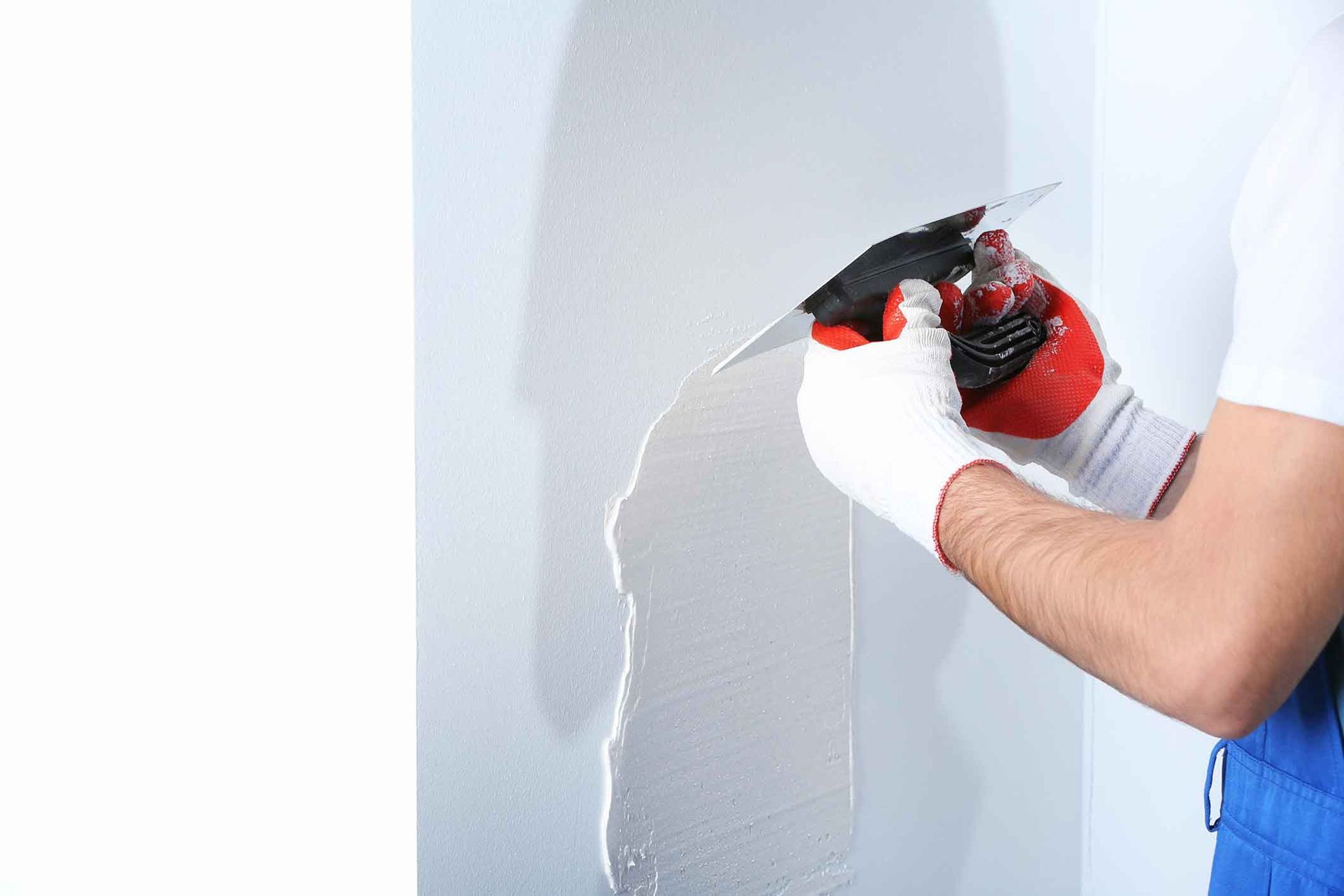 Person in blue overalls and gloves smoothing plaster on a wall with a trowel.