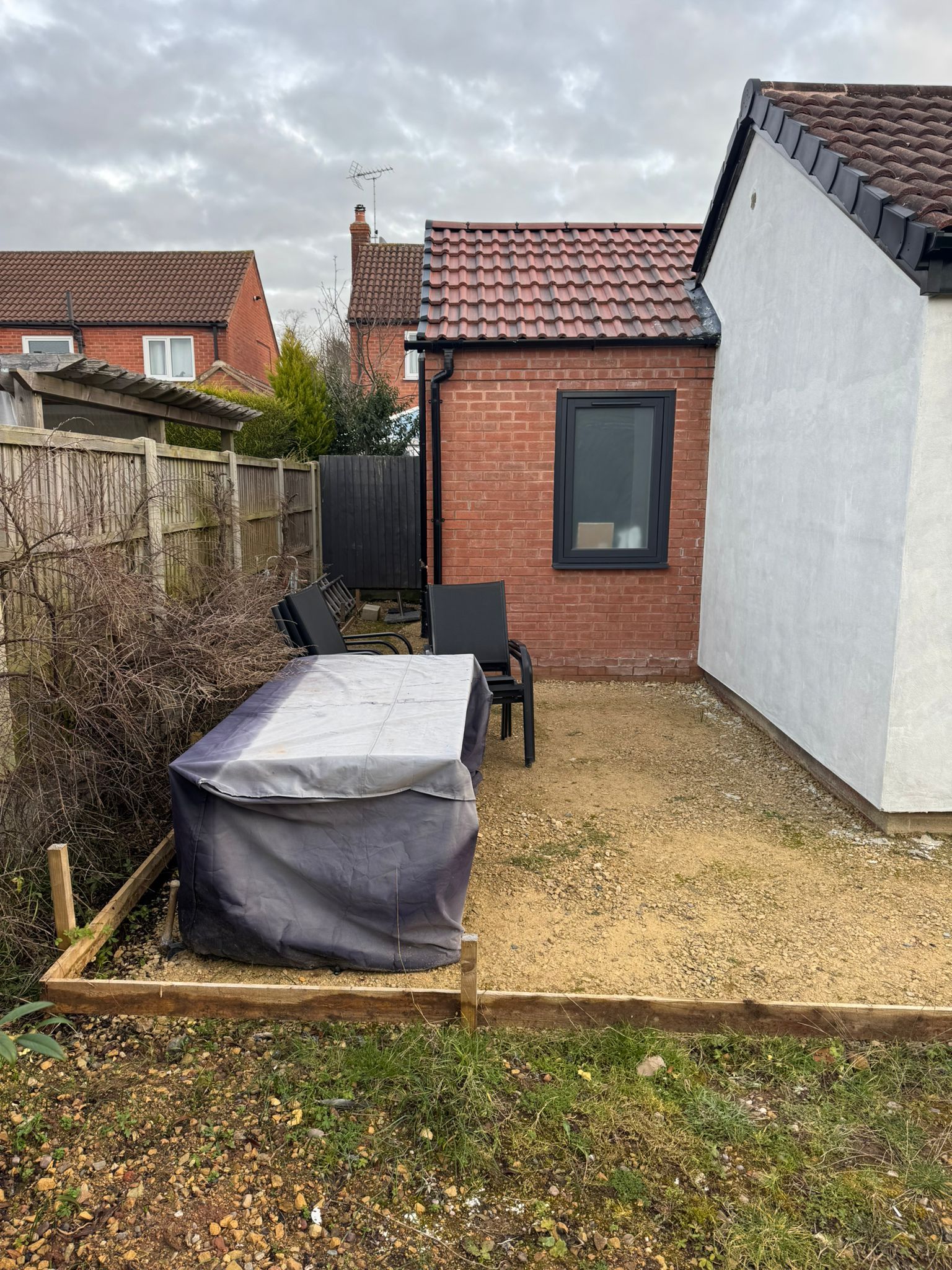 Backyard with covered table, chairs, gravel, and brick building with window. Cloudy sky.
