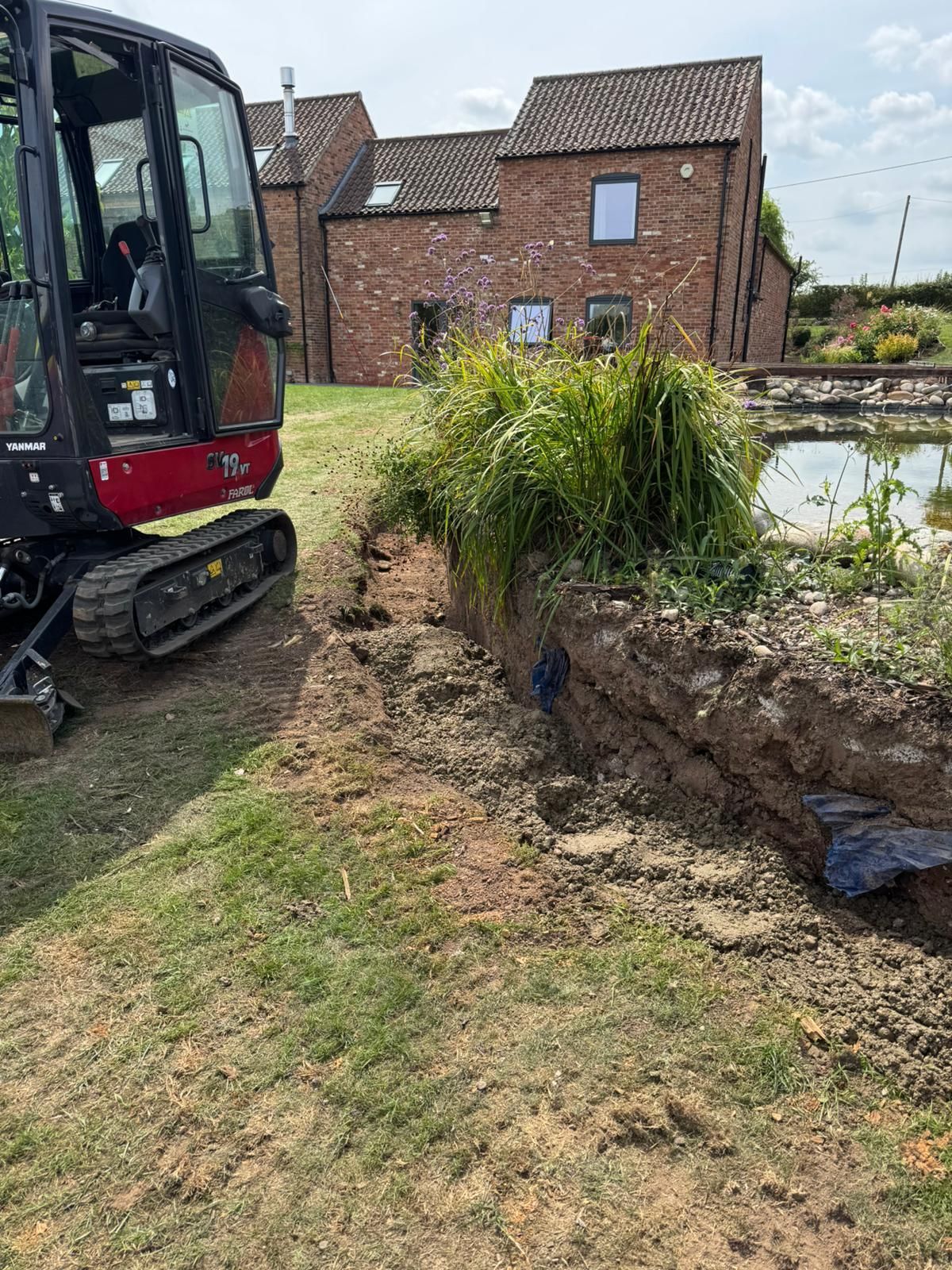 Small excavator digging trench near a pond with a brick house in the background.