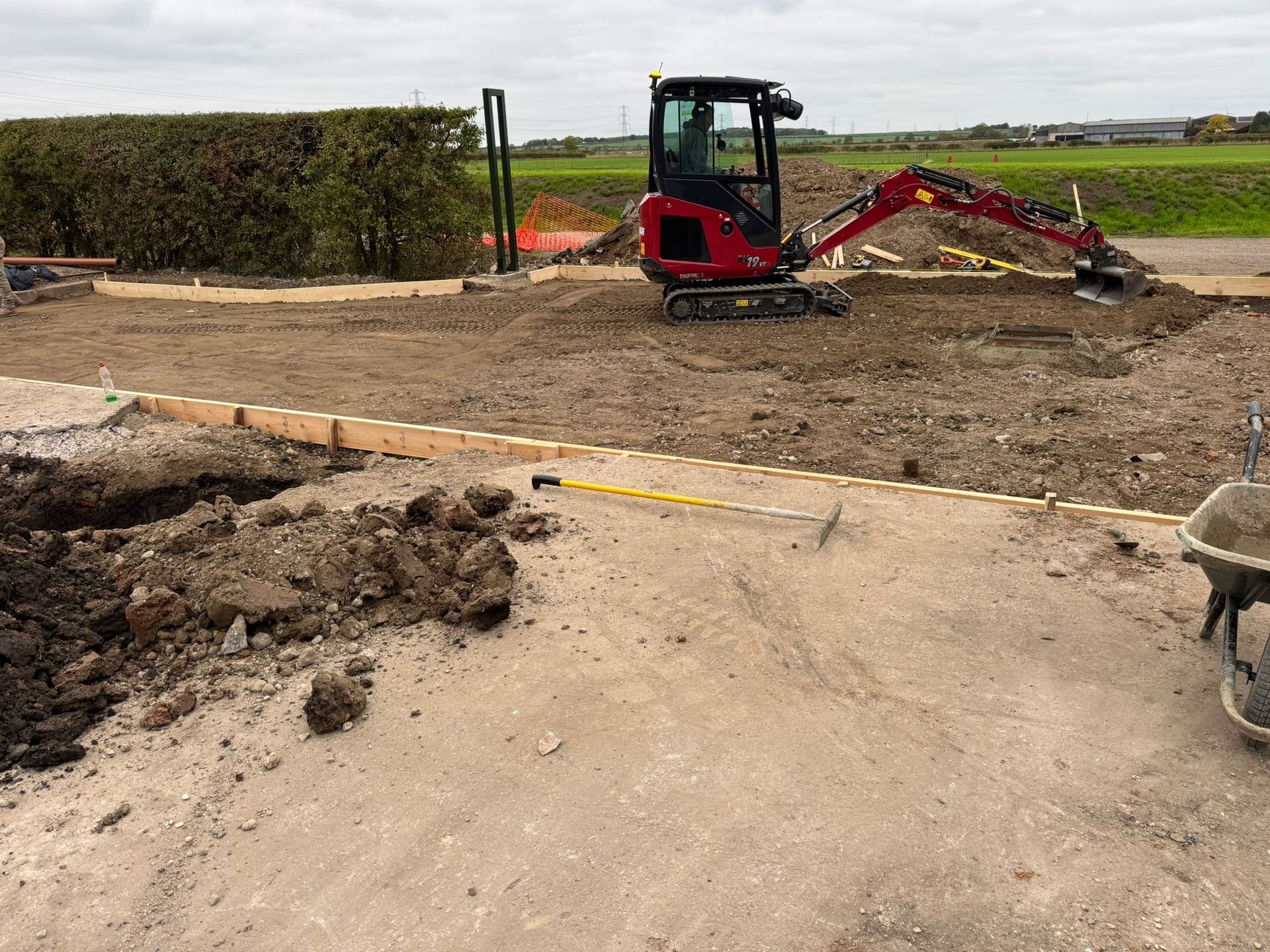 Mini excavator on a construction site, scooping dirt. Wooden border outlines a space, cloudy sky.