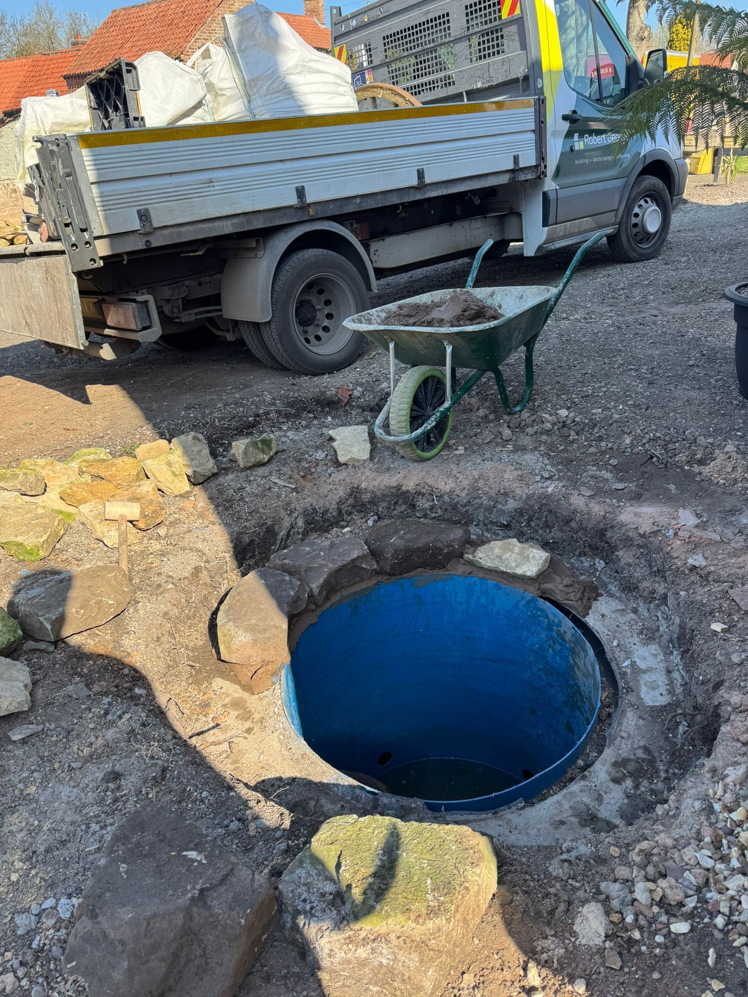 A large, open, circular hole in the ground, with a truck and wheelbarrow nearby, suggests construction.