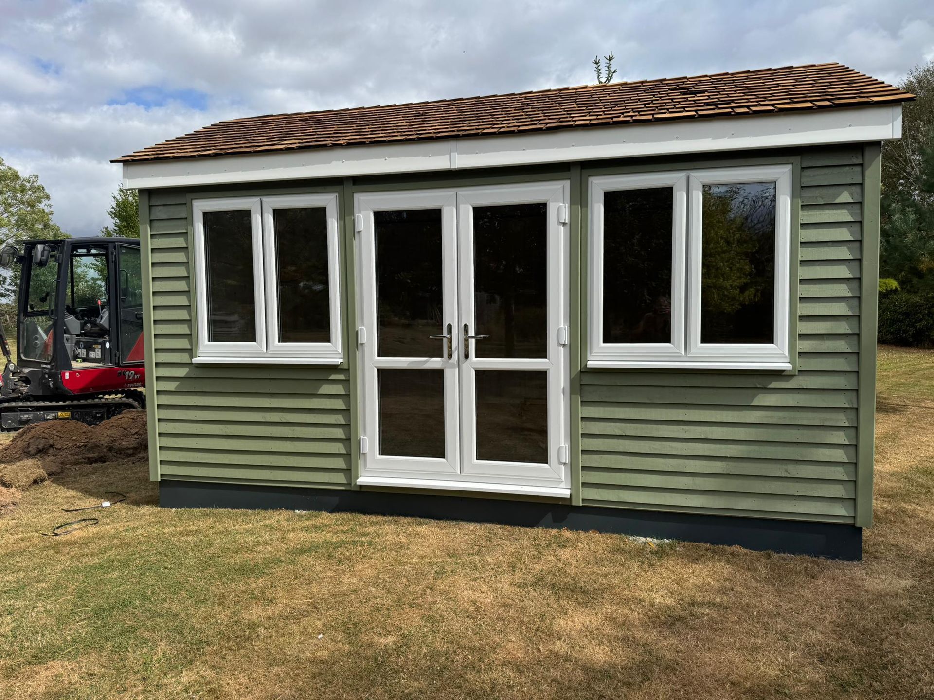Green shed with white-framed windows and doors, brown roof, set on a grassy lawn.