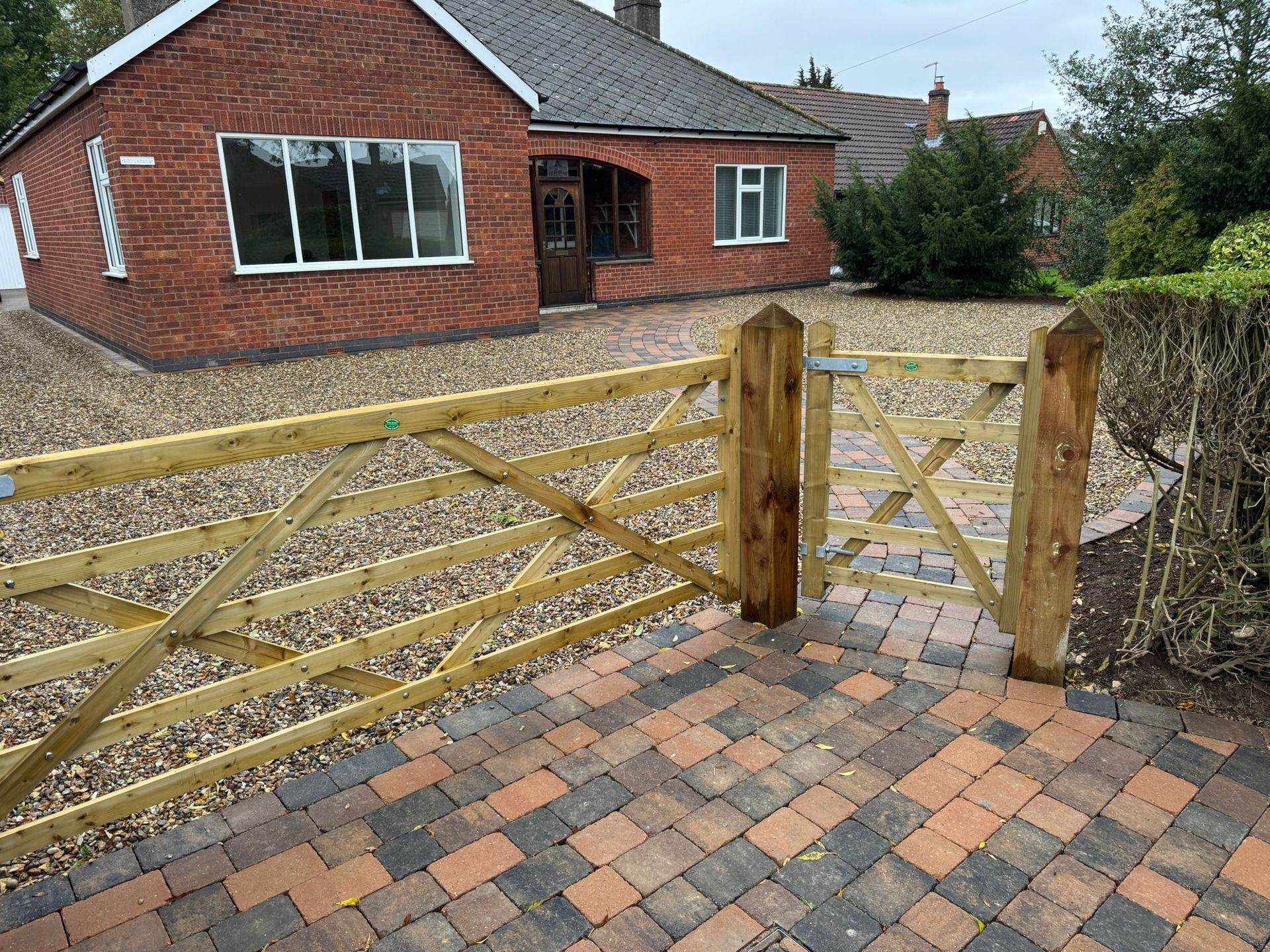 Wooden gate and fence in front of a brick house with gravel driveway and paved entrance.