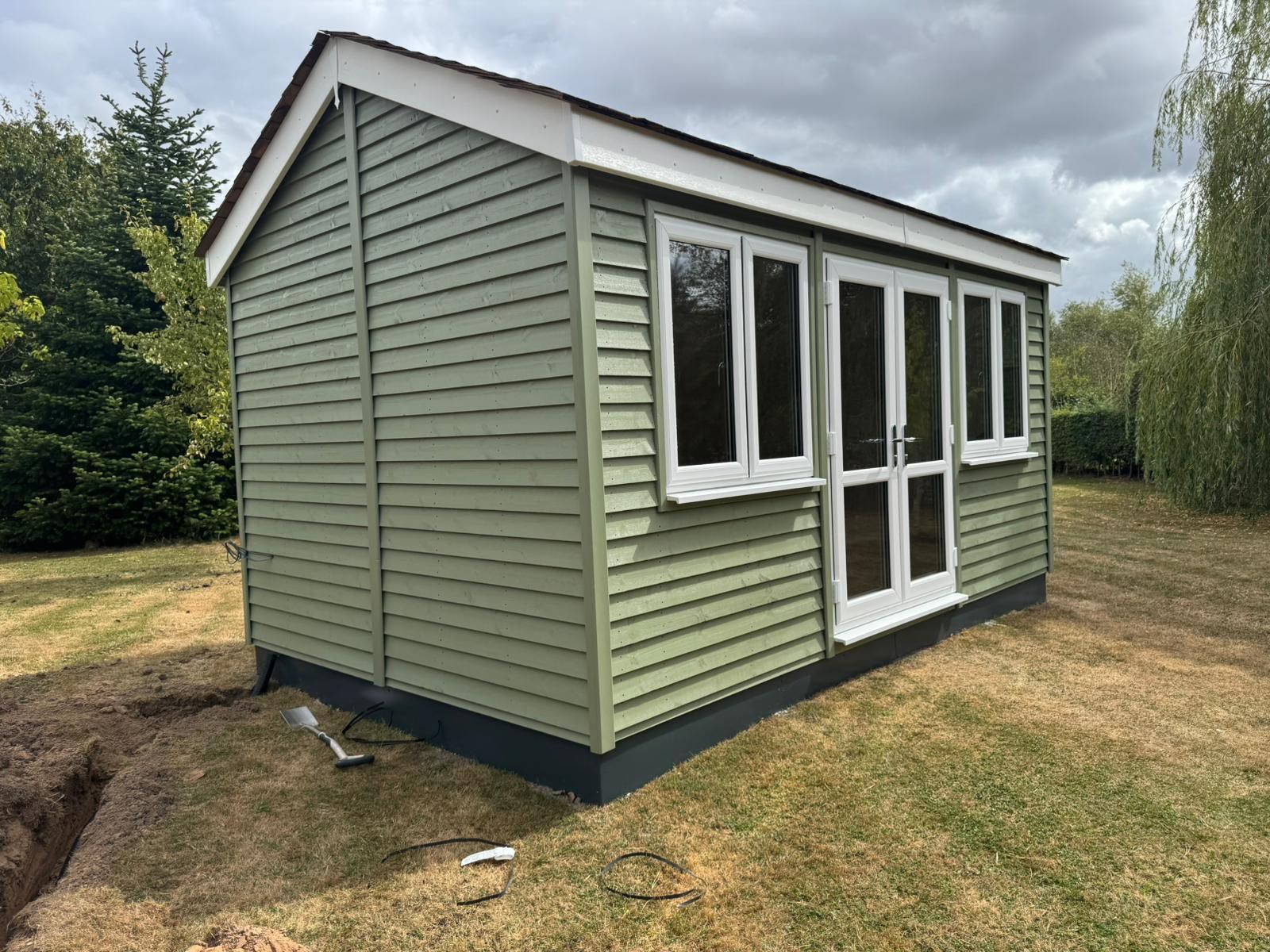 Green garden shed with white-framed windows and doors, on a grassy lawn with a small trench in front.