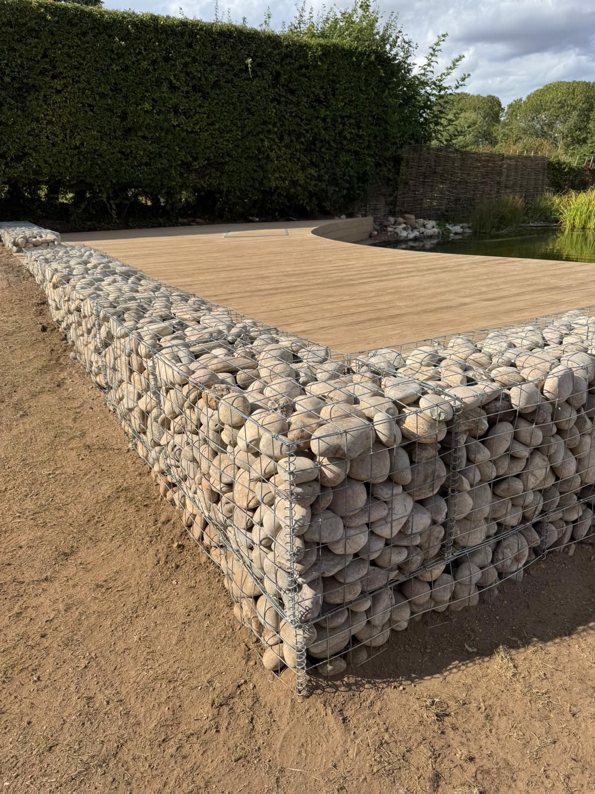 Gabion stone wall enclosing a flat, light brown, rectangular area. Green hedge in the background.