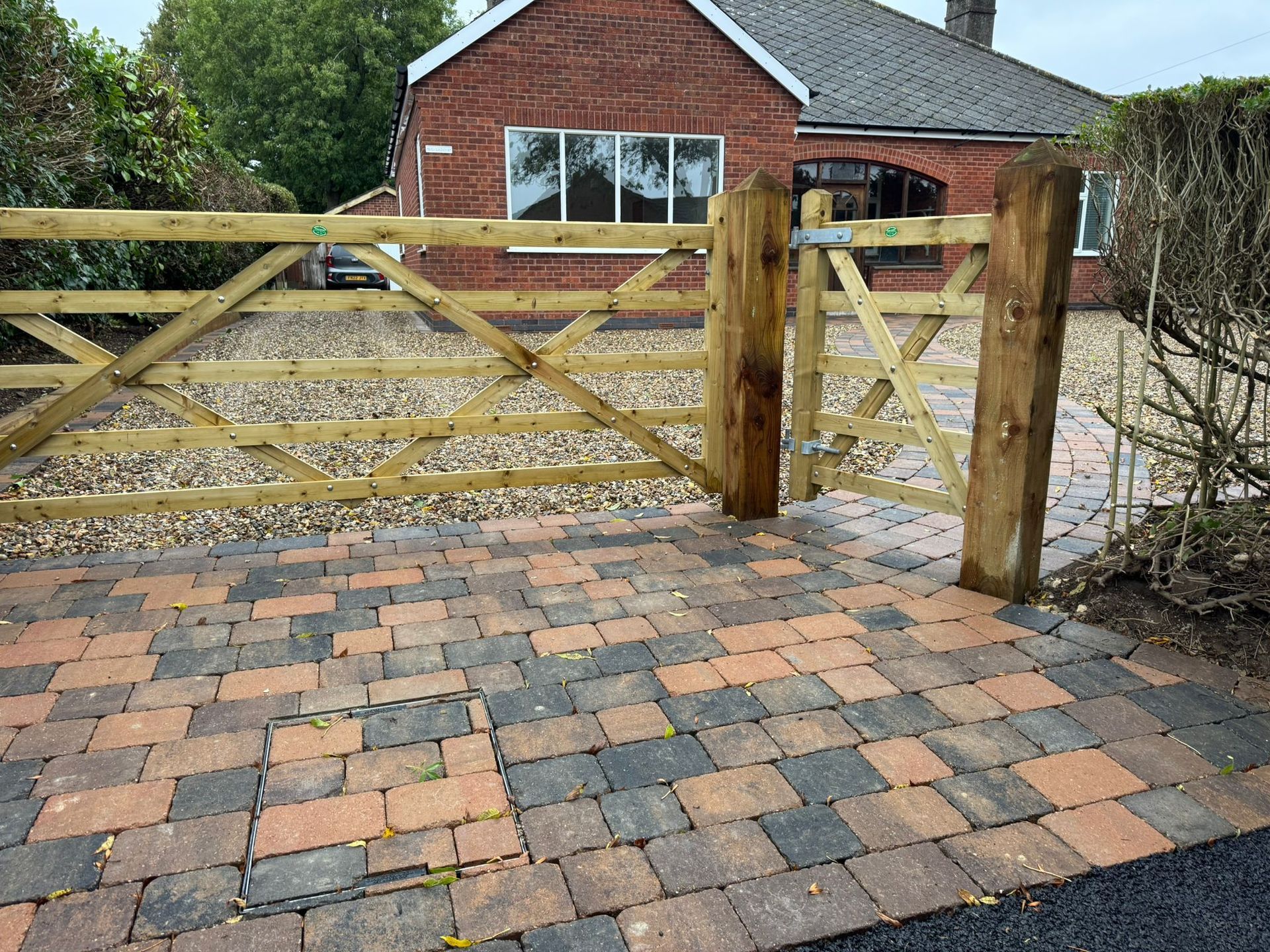 Wooden gate and posts on brick paving, leading to a gravel driveway in front of a brick house.