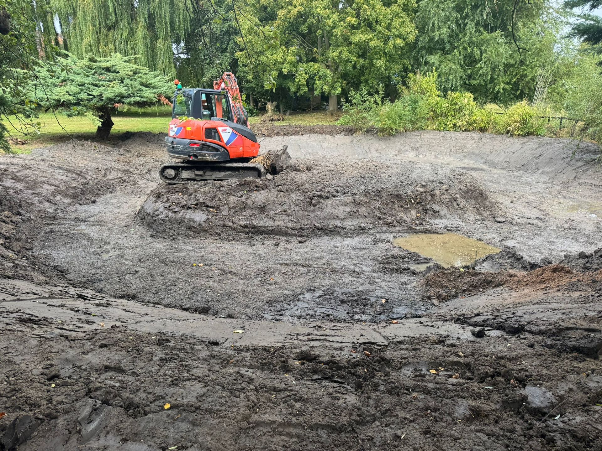 An excavator digs in a muddy pond, surrounded by trees.