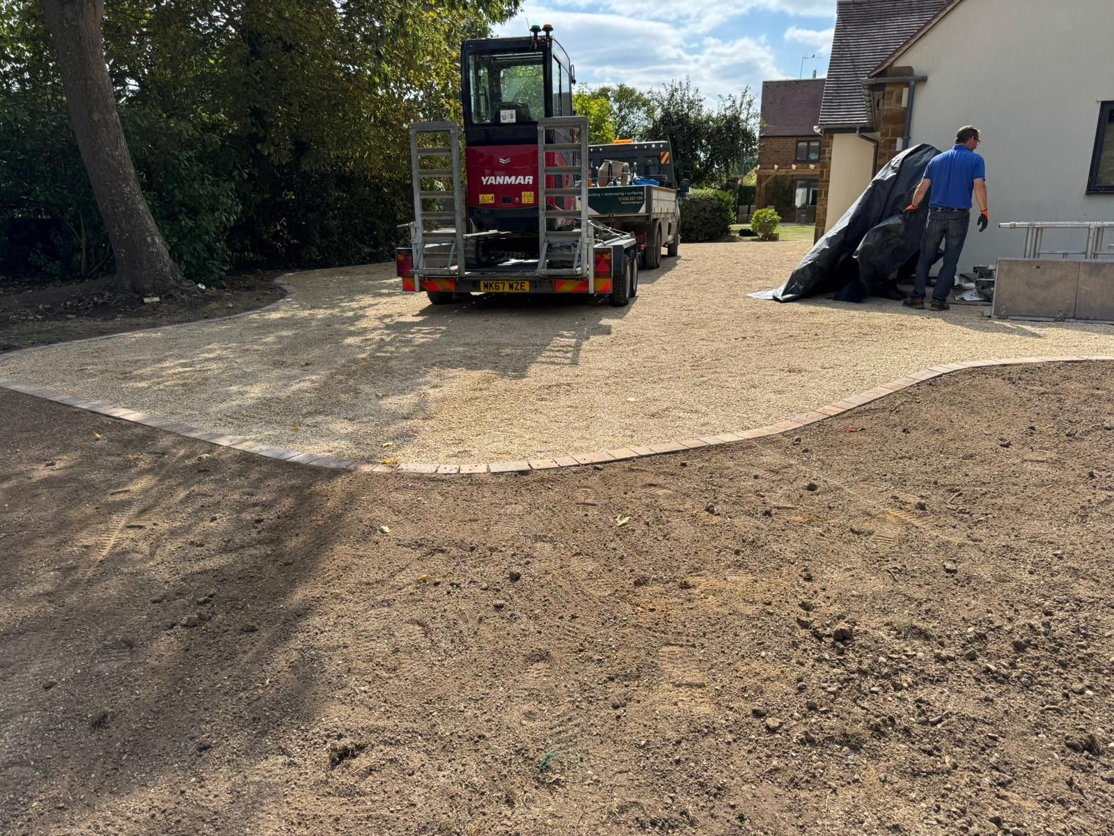 Driveway resurfacing with gravel. A mini-digger is on a trailer. A worker unloads black material.