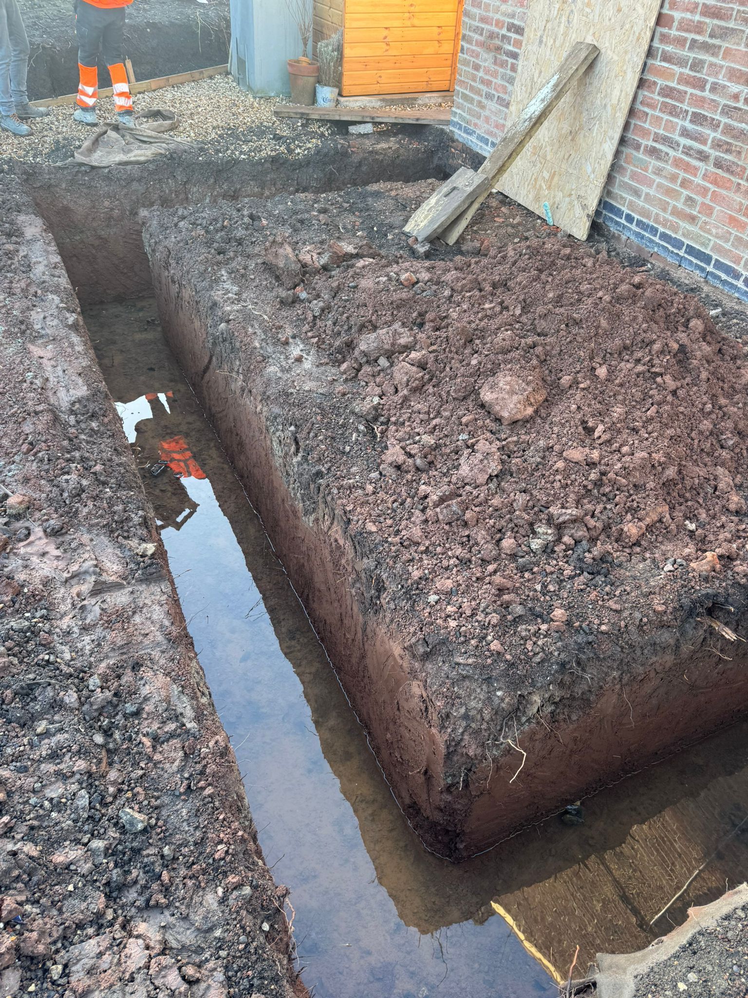 Trench filled with water, dug next to a brick building. Dirt and construction materials are scattered around.