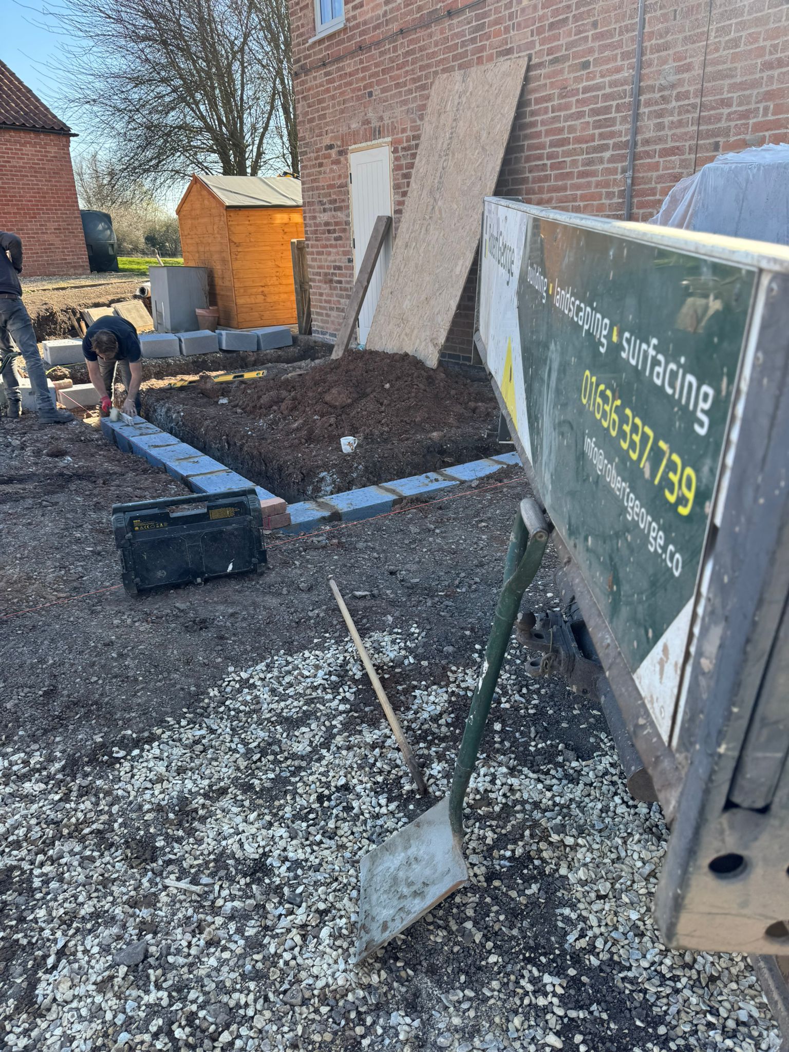 Construction site: person laying pavers next to a brick building. Shovel leans against a truck with business signage.