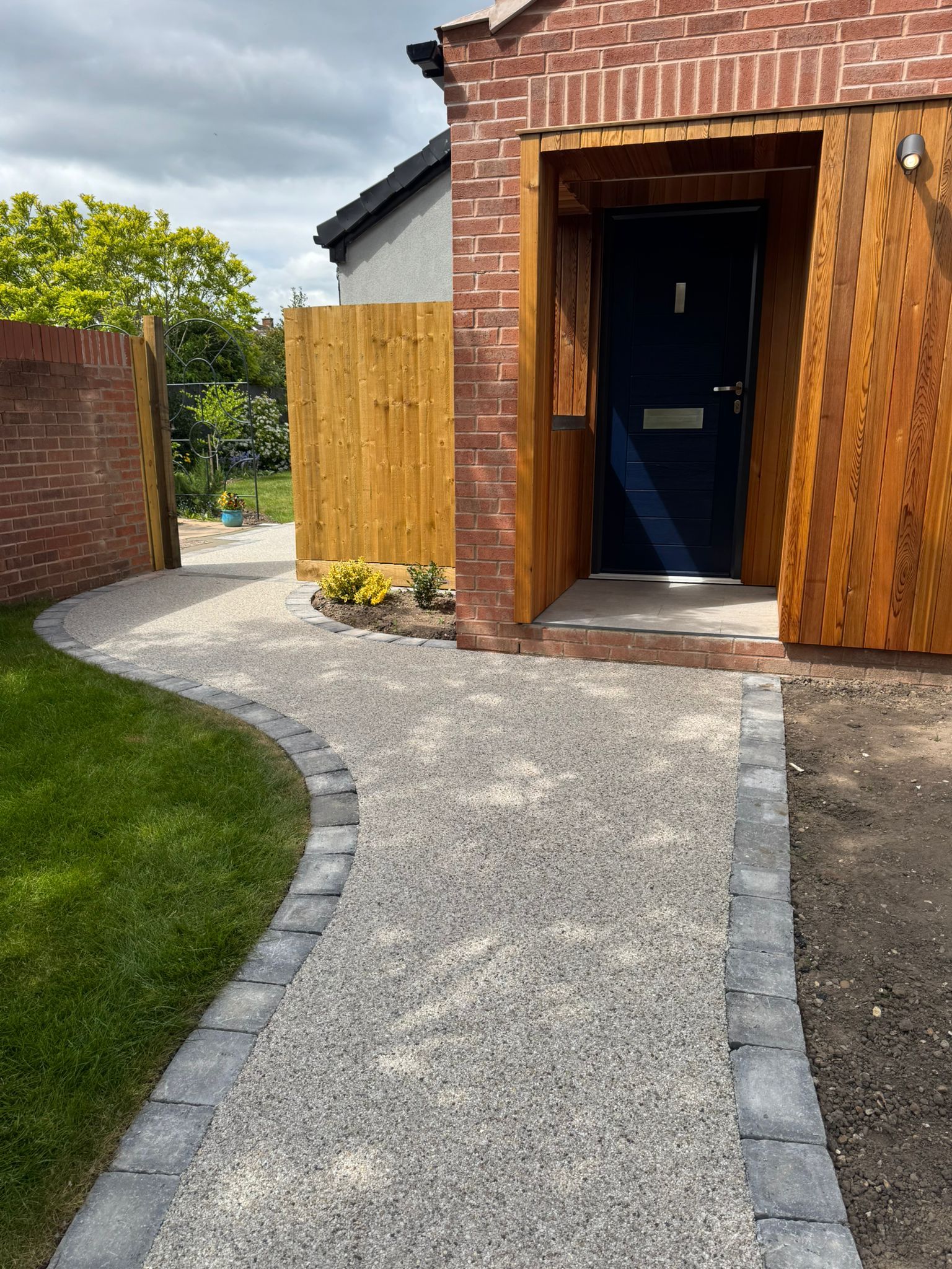A gravel pathway bordered by gray blocks leads to a navy blue door framed by red brick and wood siding.