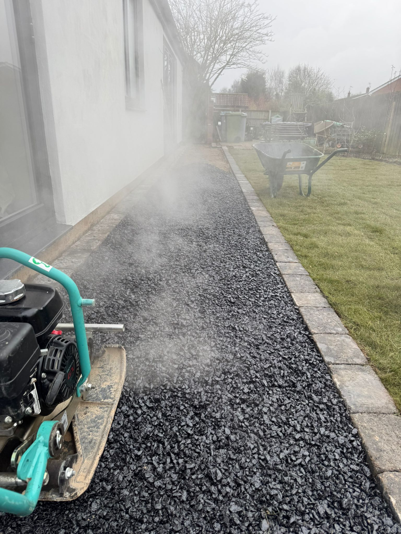 A compactor is used on a newly paved walkway. The black gravel walkway has a stone border, next to a building and lawn.