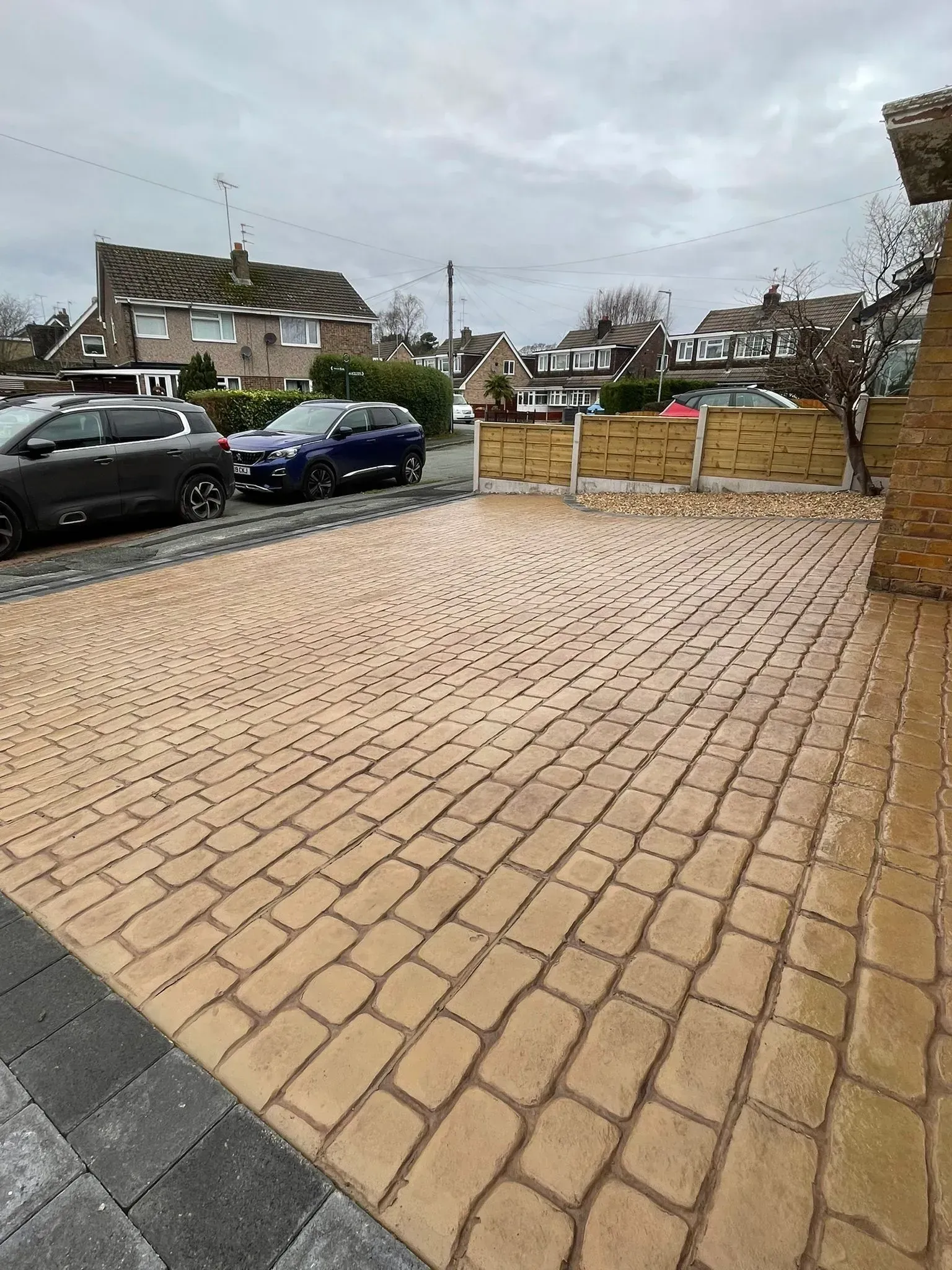 Driveway paved with tan-colored stones, bordered by gray and black pavers; cars parked on the street.
