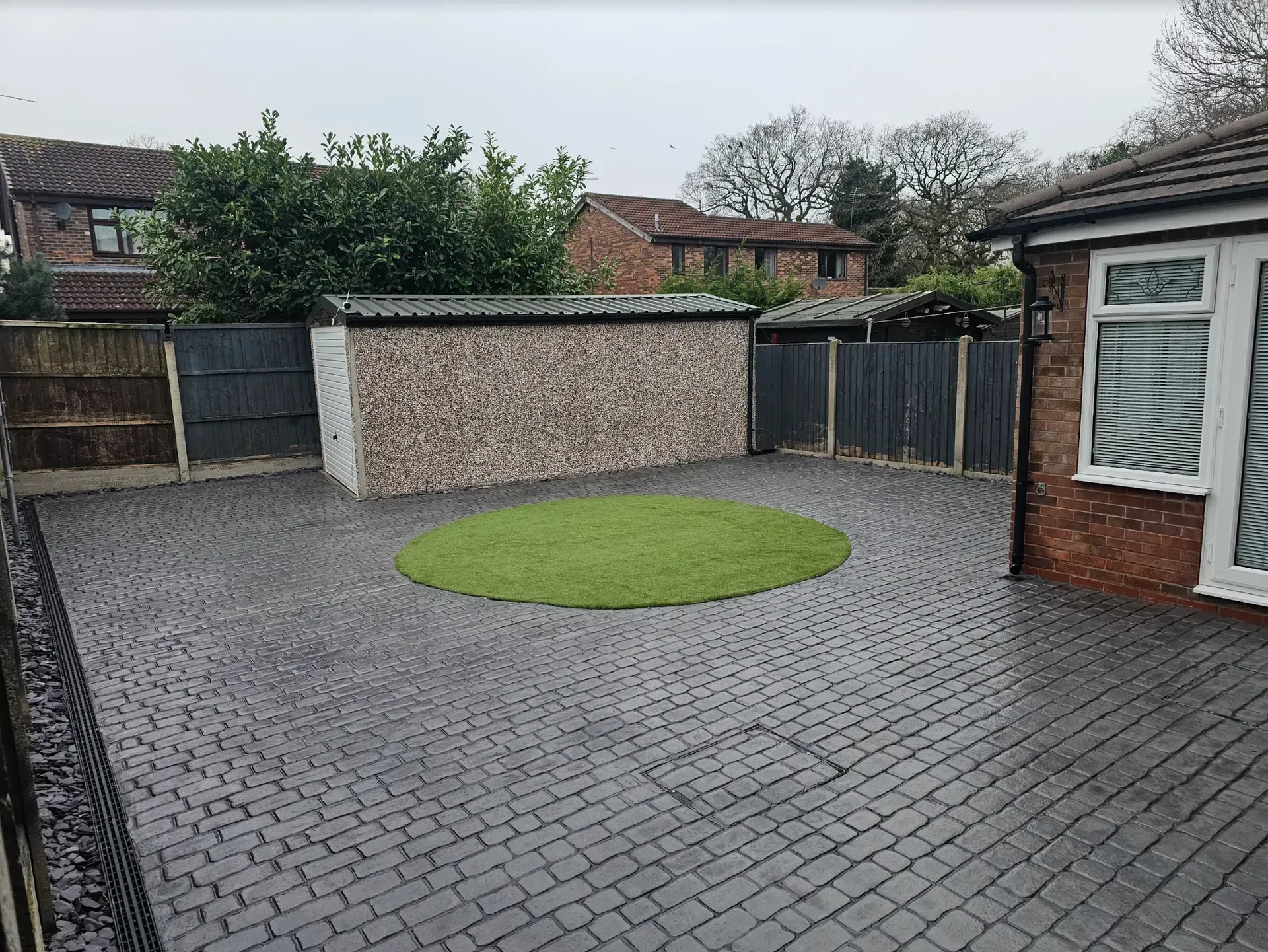 Gray brick driveway with a circular patch of green grass; a stone shed and fence.
