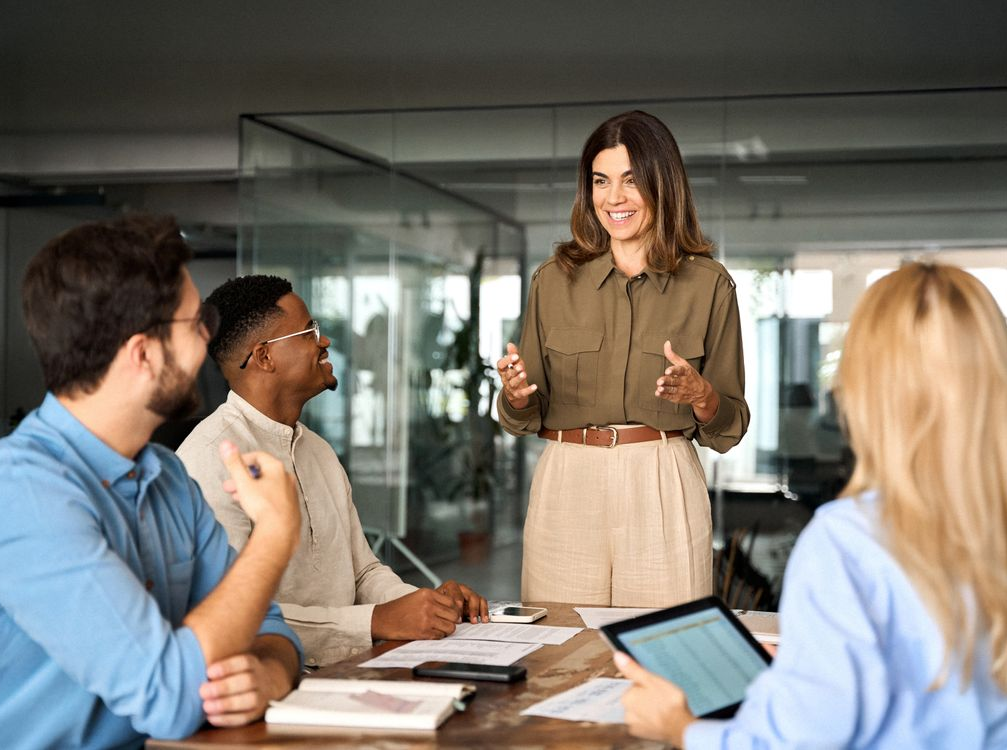 Business meeting with three people listening to a woman presenting in a glass-walled office