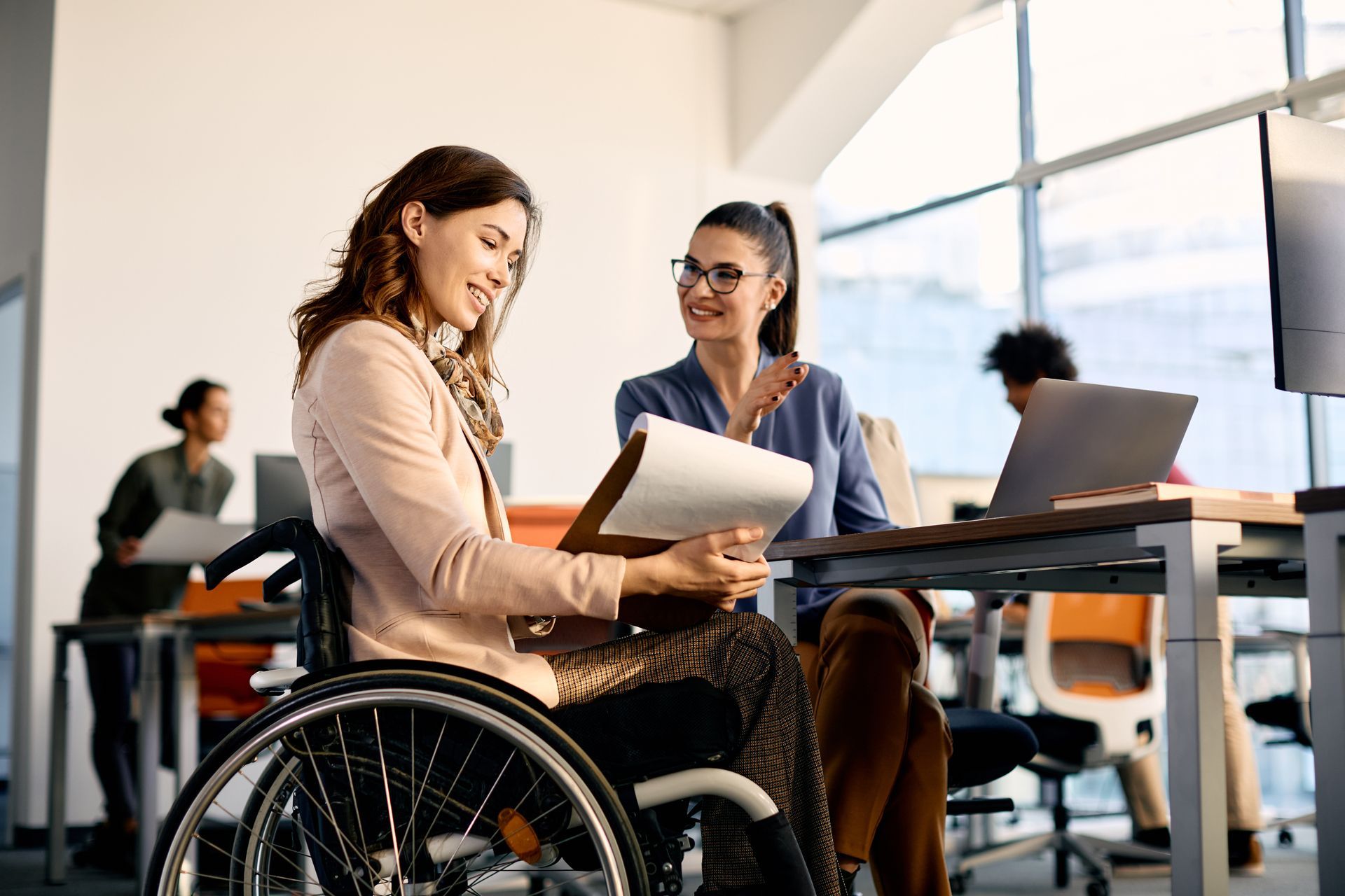 Woman in wheelchair reviews documents with colleague at an office desk.