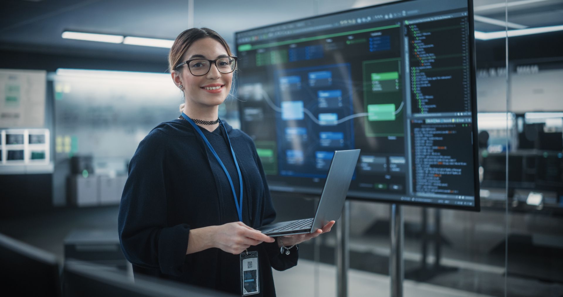 Person with glasses using a laptop in a modern control room with large data displays