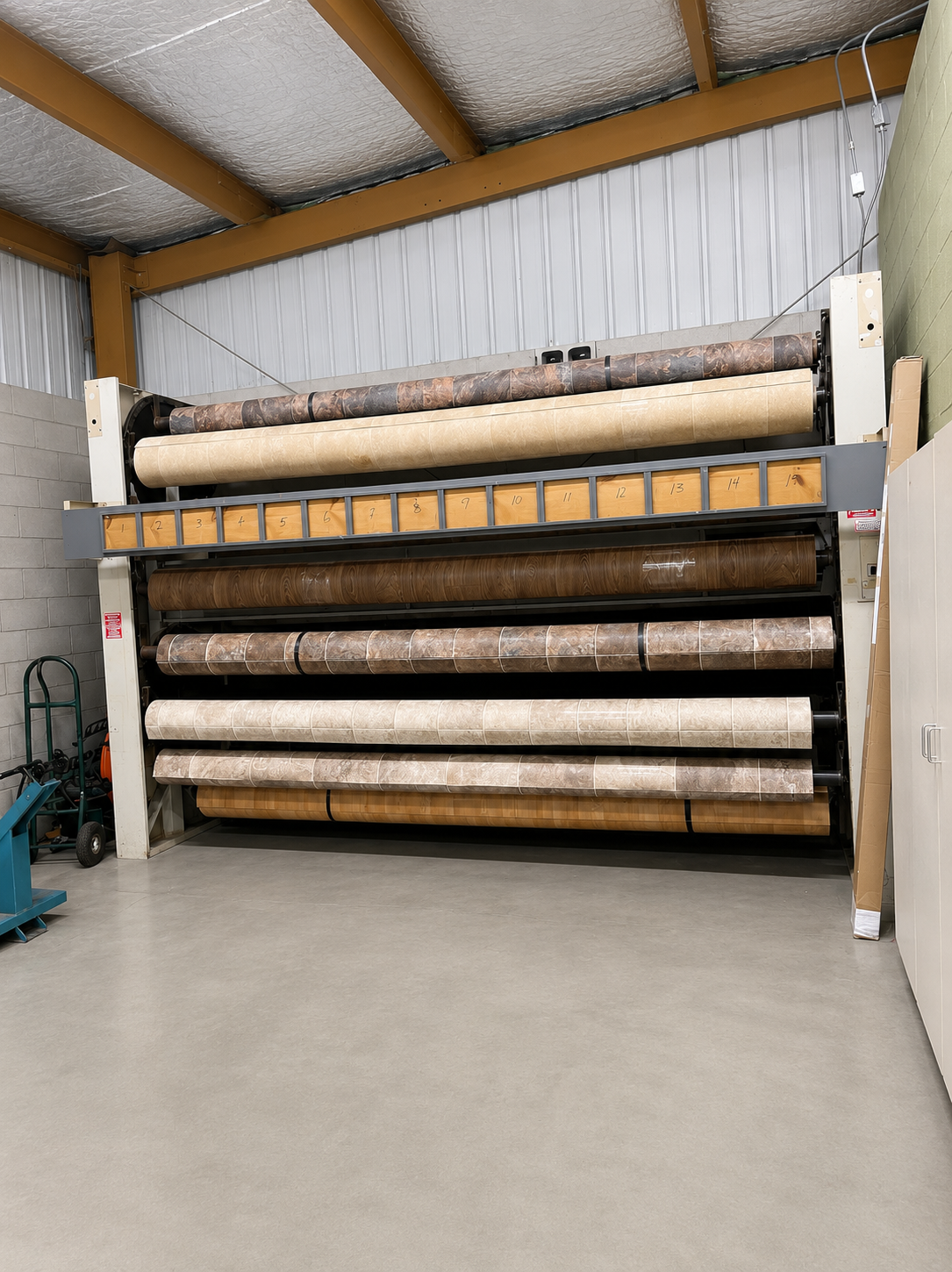 Rolls of carpet stacked in a warehouse on a metal rack under a corrugated roof