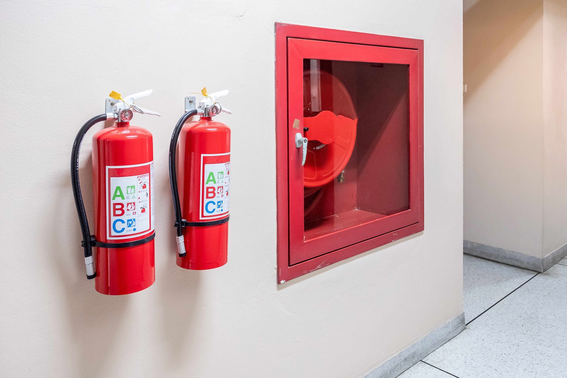 Two red fire extinguishers beside a wall-mounted fire hose cabinet in a hallway Two red fire extinguishers beside a wall-mounted fire hose cabinet in a hallway
