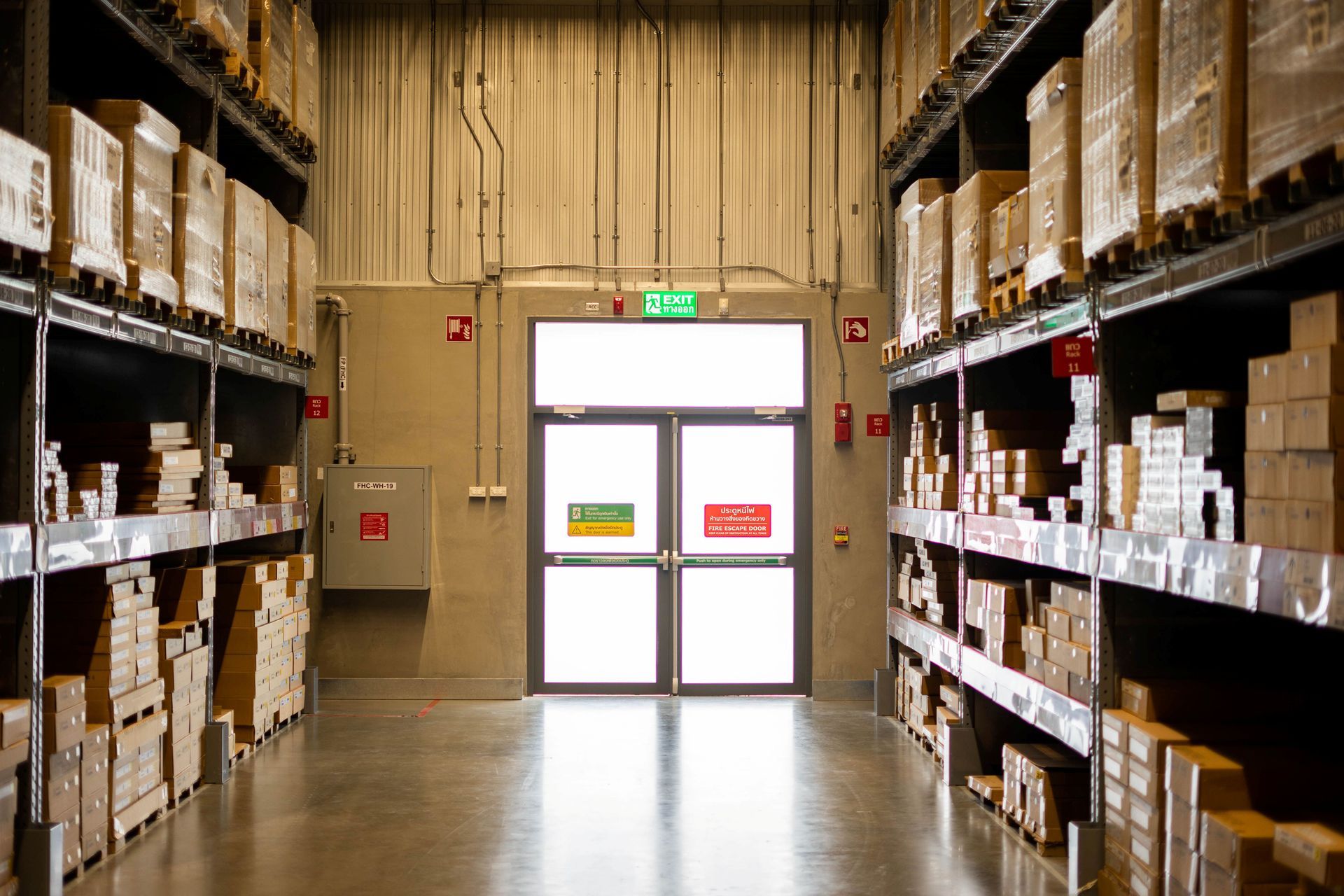 Warehouse aisle with stacked boxes and shelves leading to a closed double door in the center.