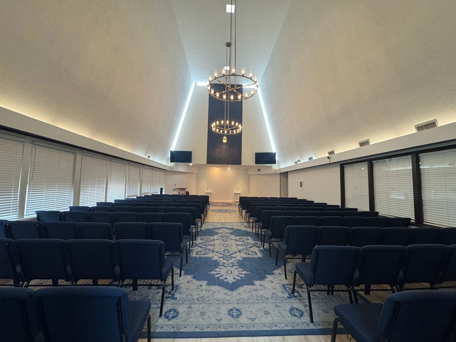 Empty conference room with rows of navy chairs facing a central stage and chandelier under a vaulted ceiling
