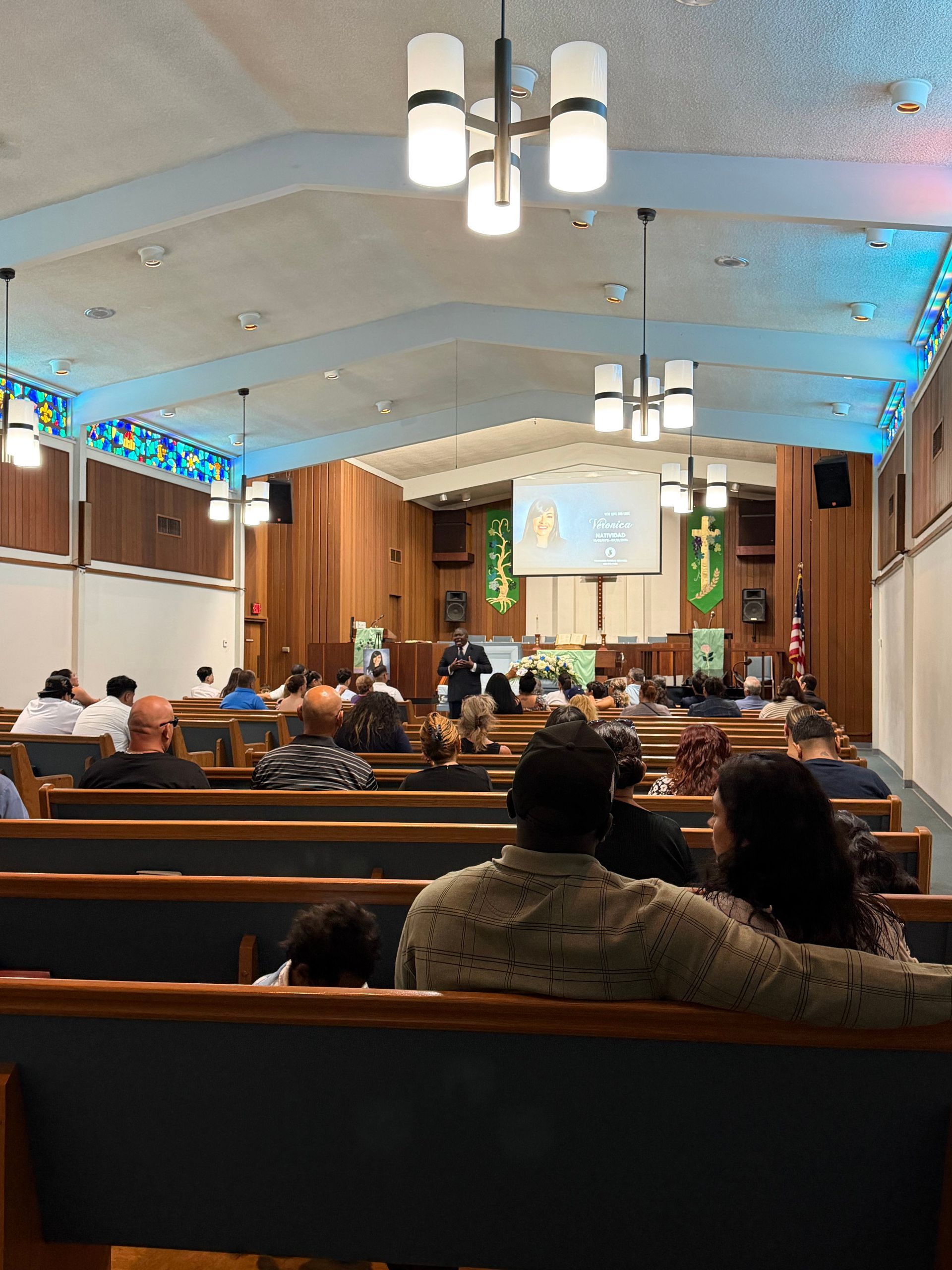 Church sanctuary with people seated in pews facing a stage and projection screen during a service