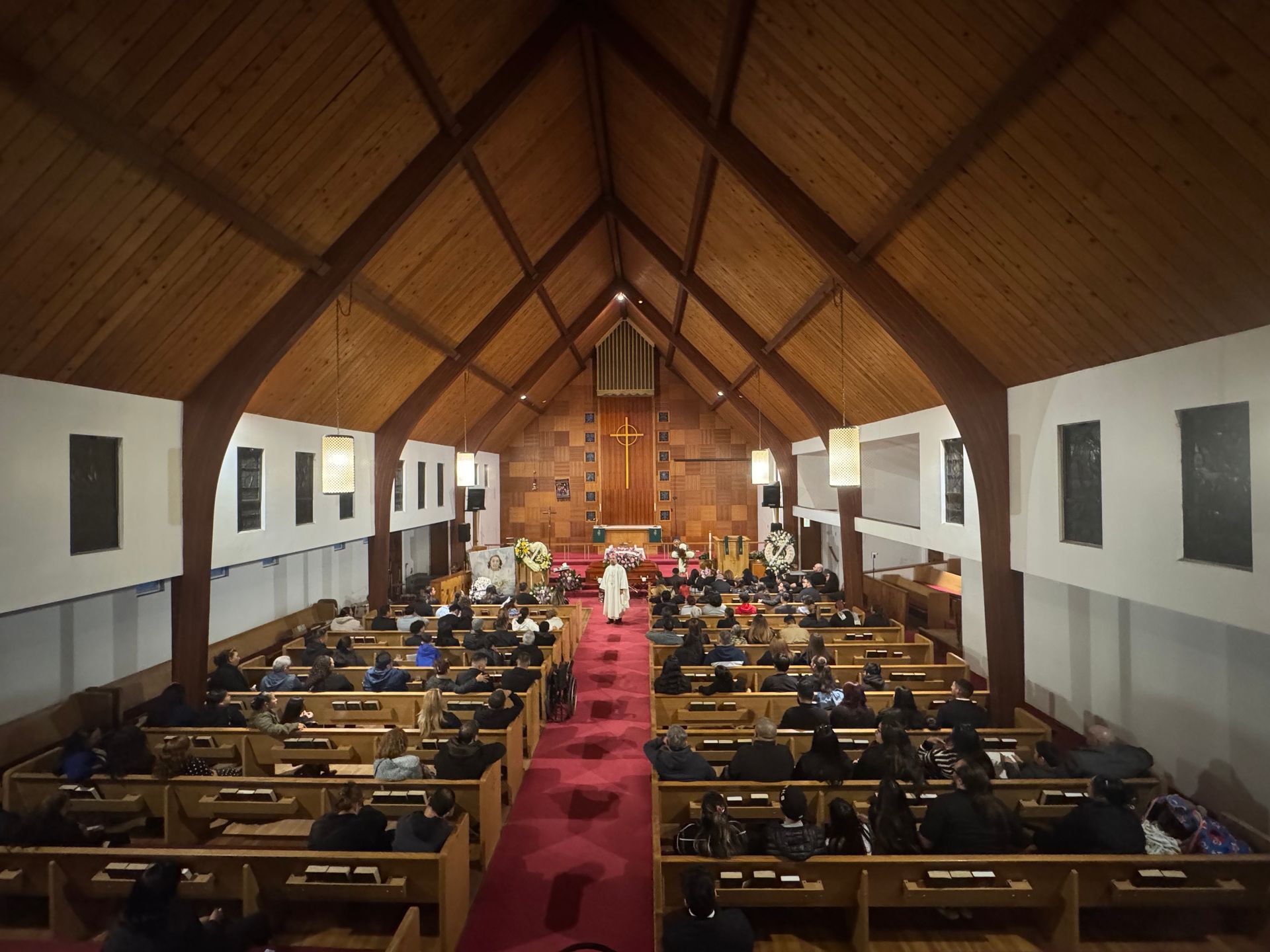 Wood-paneled church interior with people seated in pews facing the altar, red carpet down the center.