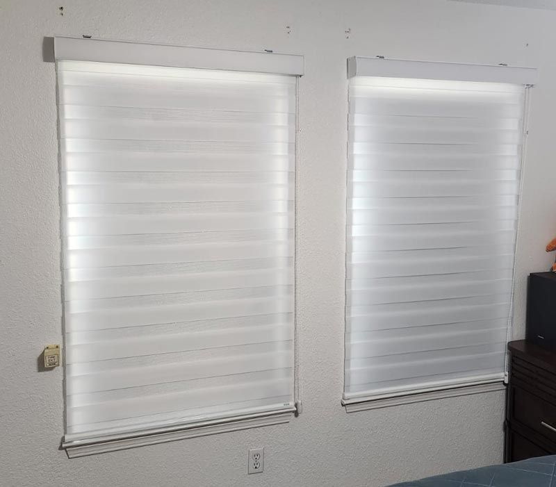 Two white window blinds on a light gray wall, with a small dark cabinet at right.