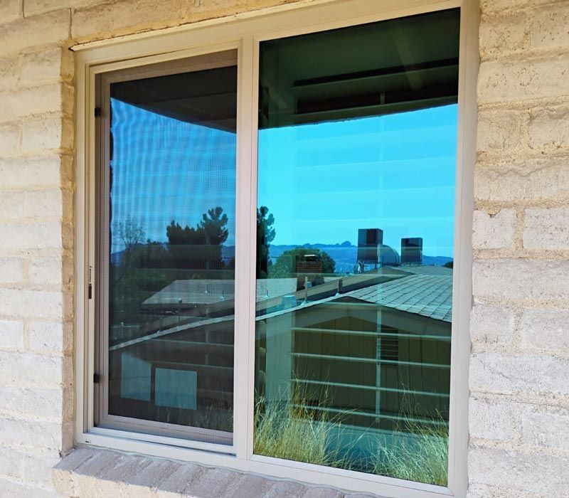 Sliding window with beige stone frame reflecting rooftops and blue sky, with blinds partly visible inside