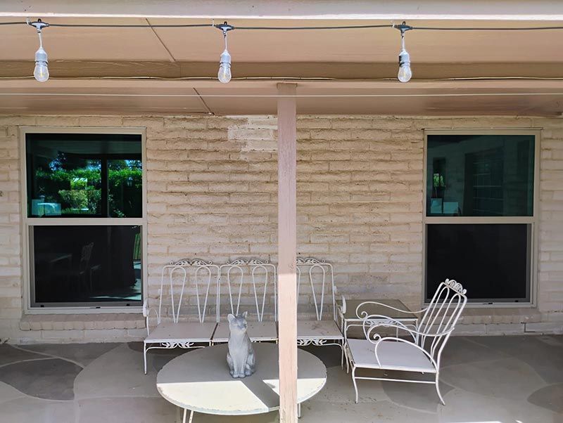Patio with white chairs and round table against a beige brick wall, under hanging lights.