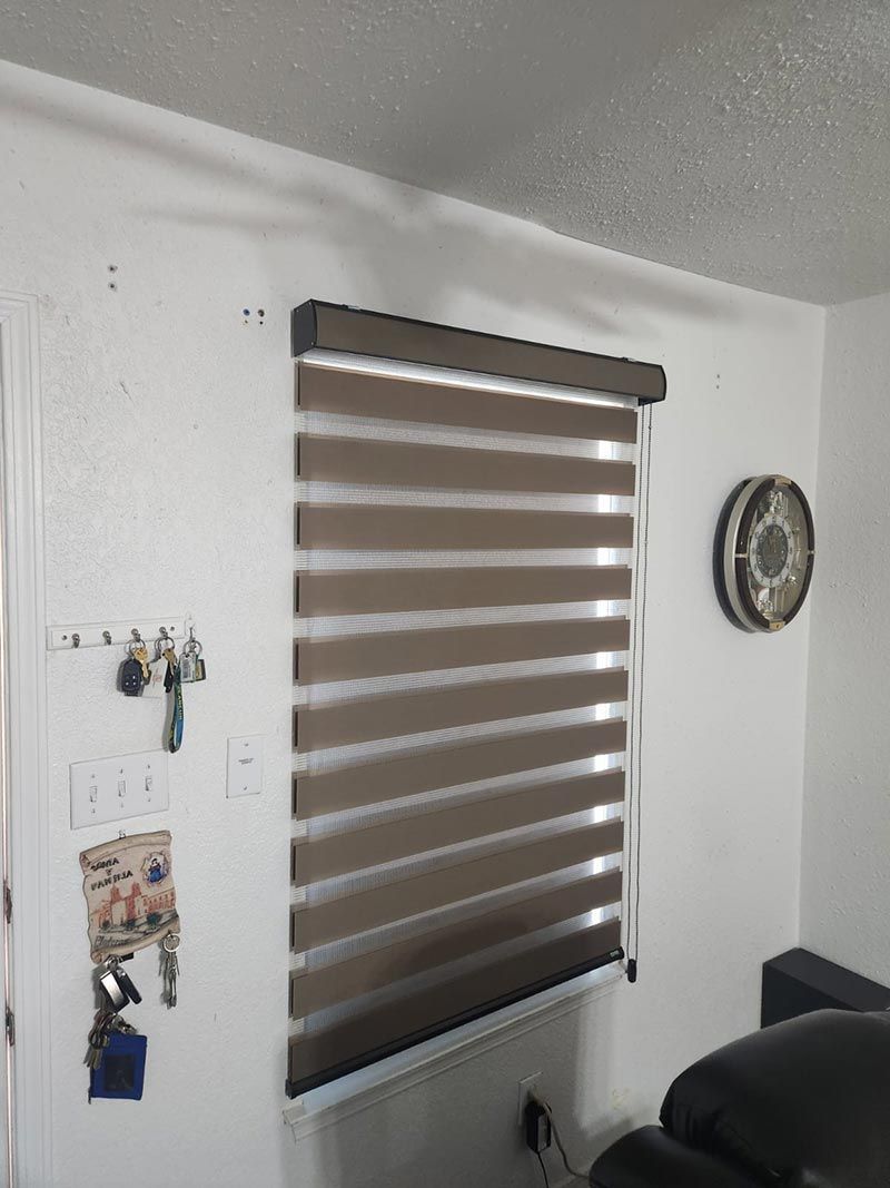 Window with brown-and-white striped blinds, wall decor, and part of a black chair in a white room