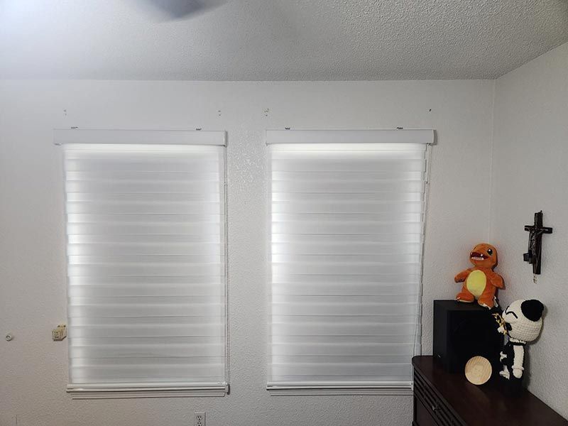 White wall with two windows covered by blinds, a black cabinet, stuffed toys, and a wall cross.