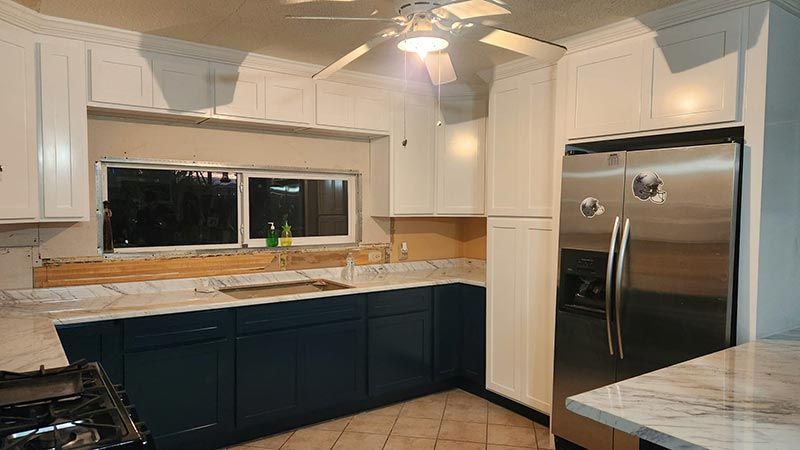 Modern kitchen with dark cabinets, white counters, stainless steel fridge, and a window over the sink.