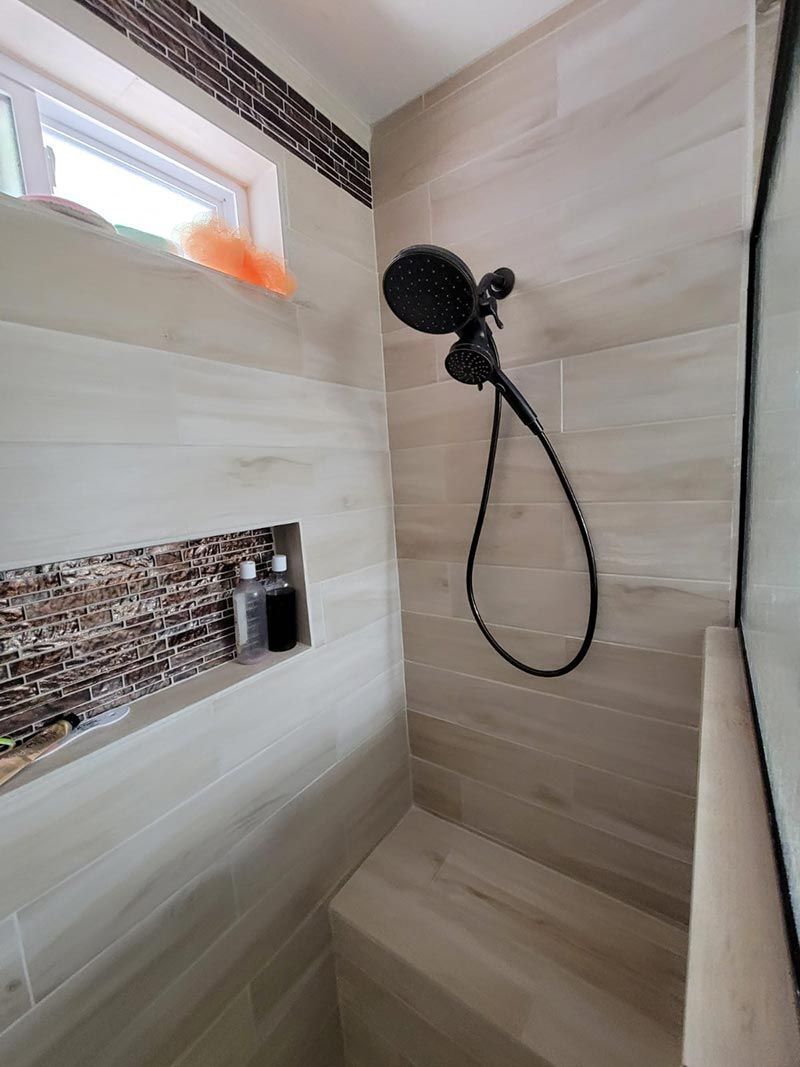Modern tiled shower with black handheld showerhead, bench, and small window shelf with orange item