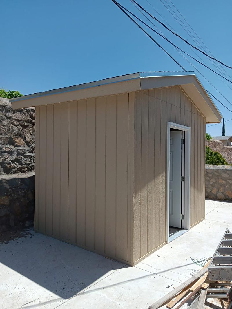 Small beige shed with an open white door on a concrete pad beside a stone wall under a blue sky