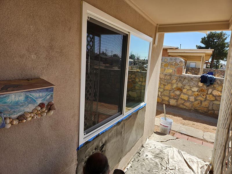Narrow outdoor walkway beside a stucco house, with a window, paint bucket, and stone wall in sunlight