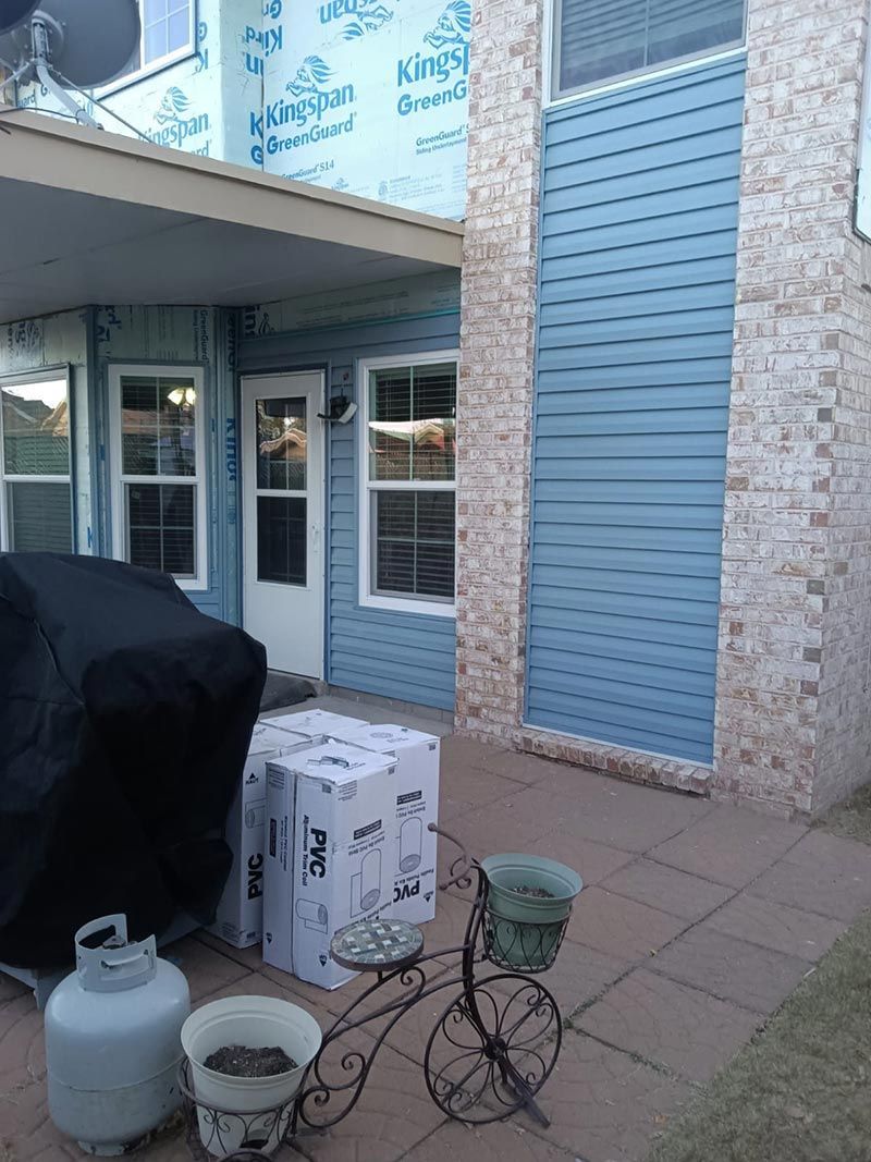 Patio with covered grill, propane tank, flower pot, bicycle, and cardboard box beside a brick house