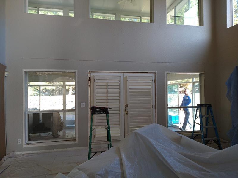 Unfinished living room with tall windows, covered furniture, ladders, and a worker near a doorway.