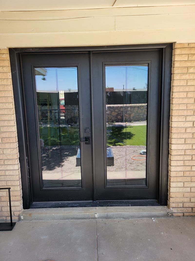 Black double glass doors set in a tan brick wall, reflecting an outdoor courtyard.