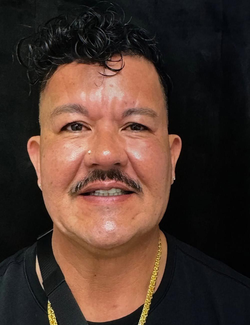 Close-up portrait of a smiling man with curly hair, mustache, and gold necklace against a dark background