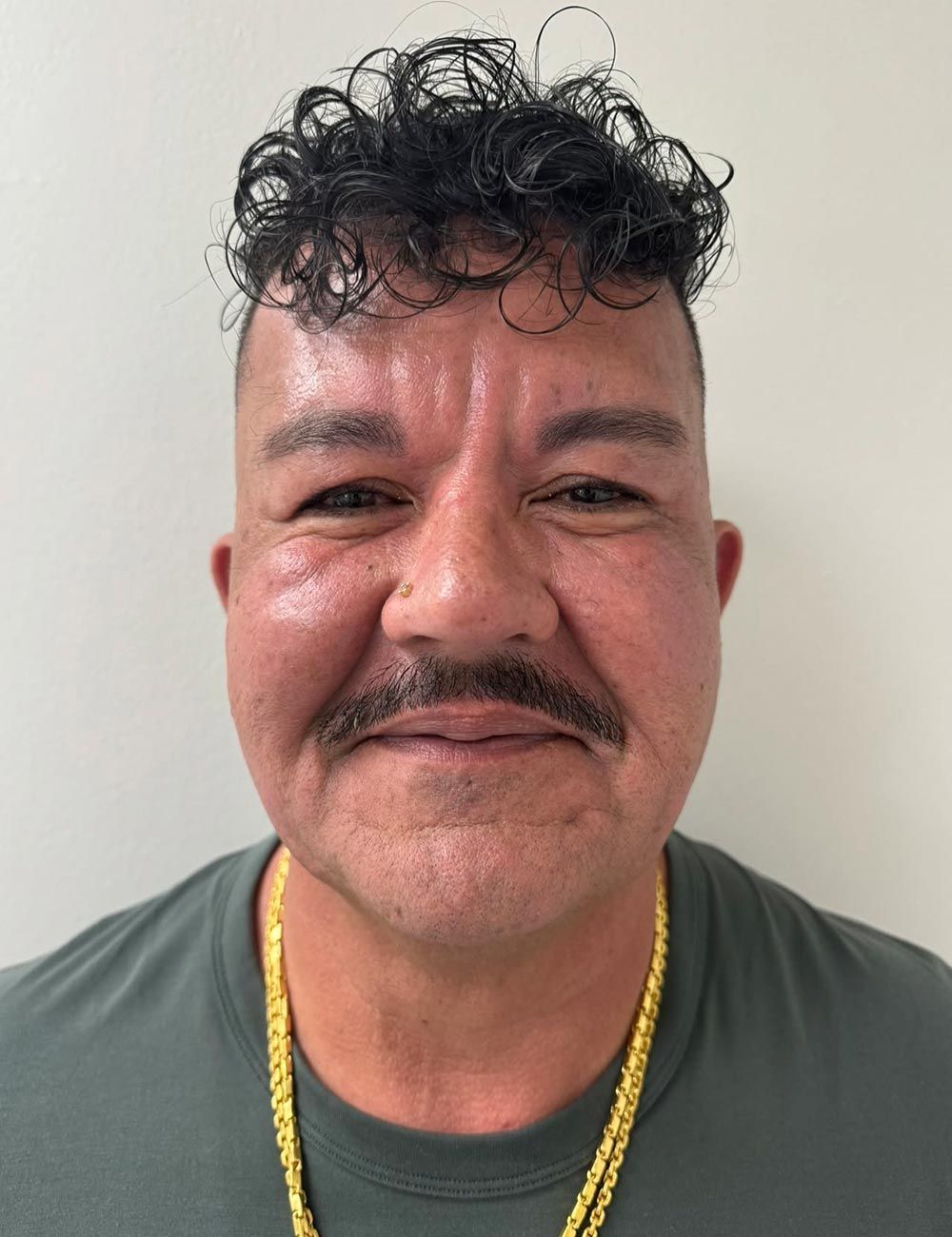 Smiling man with curly hair and a mustache wearing a gray shirt and gold chain against a plain wall