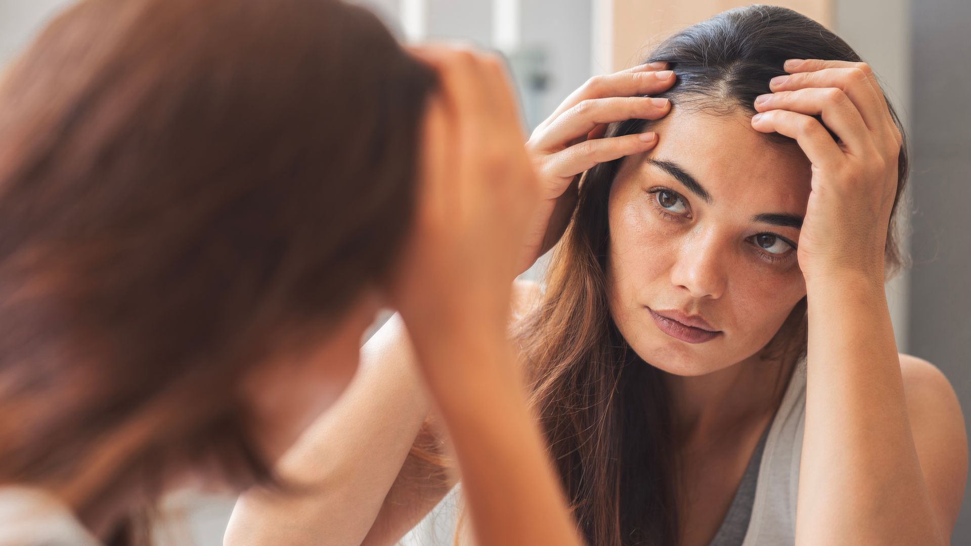 Woman looking in mirror and touching her hairline, checking for hair loss or scalp changes.