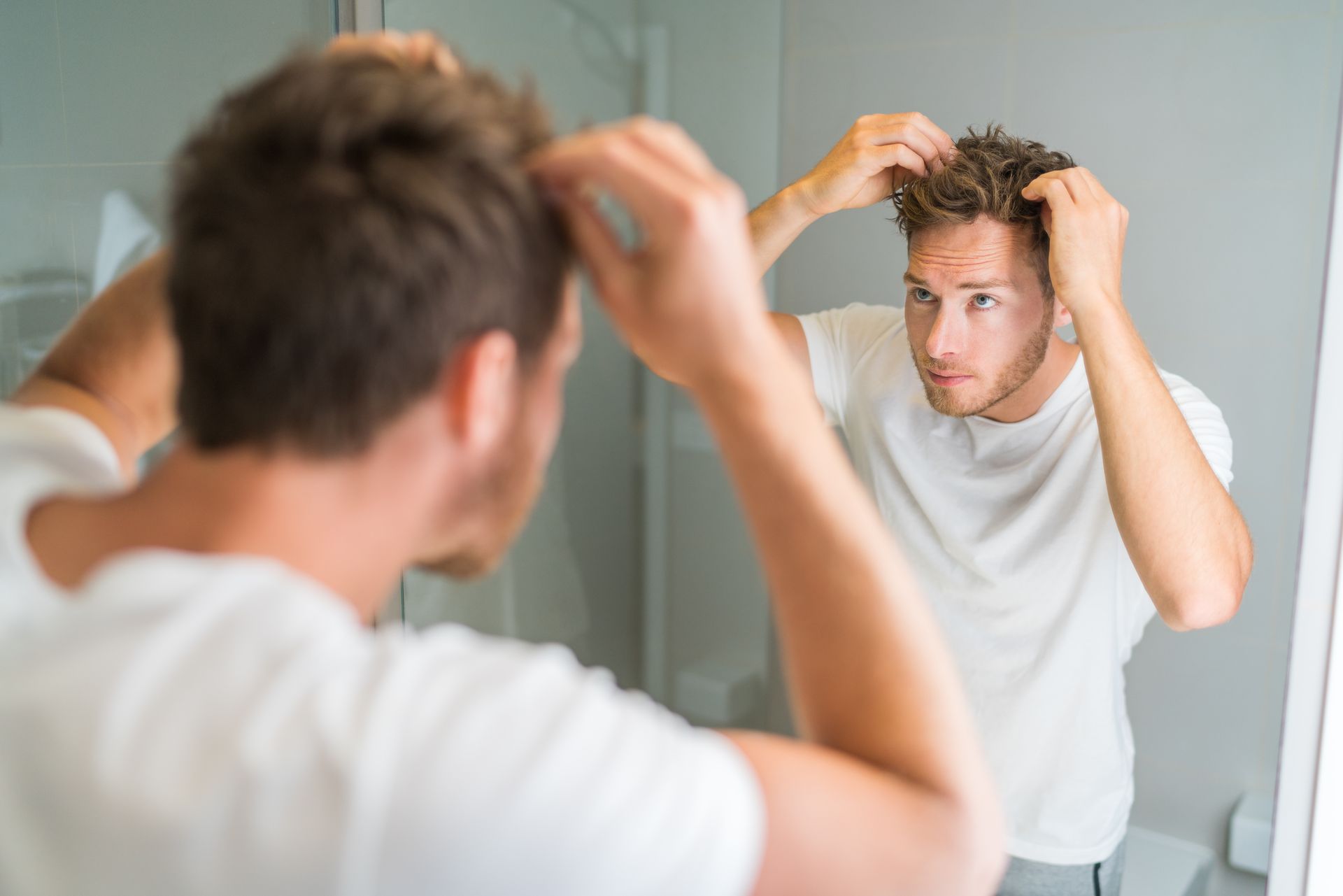 Man adjusting his hair in a bathroom mirror, wearing a white shirt, with a focused expression.