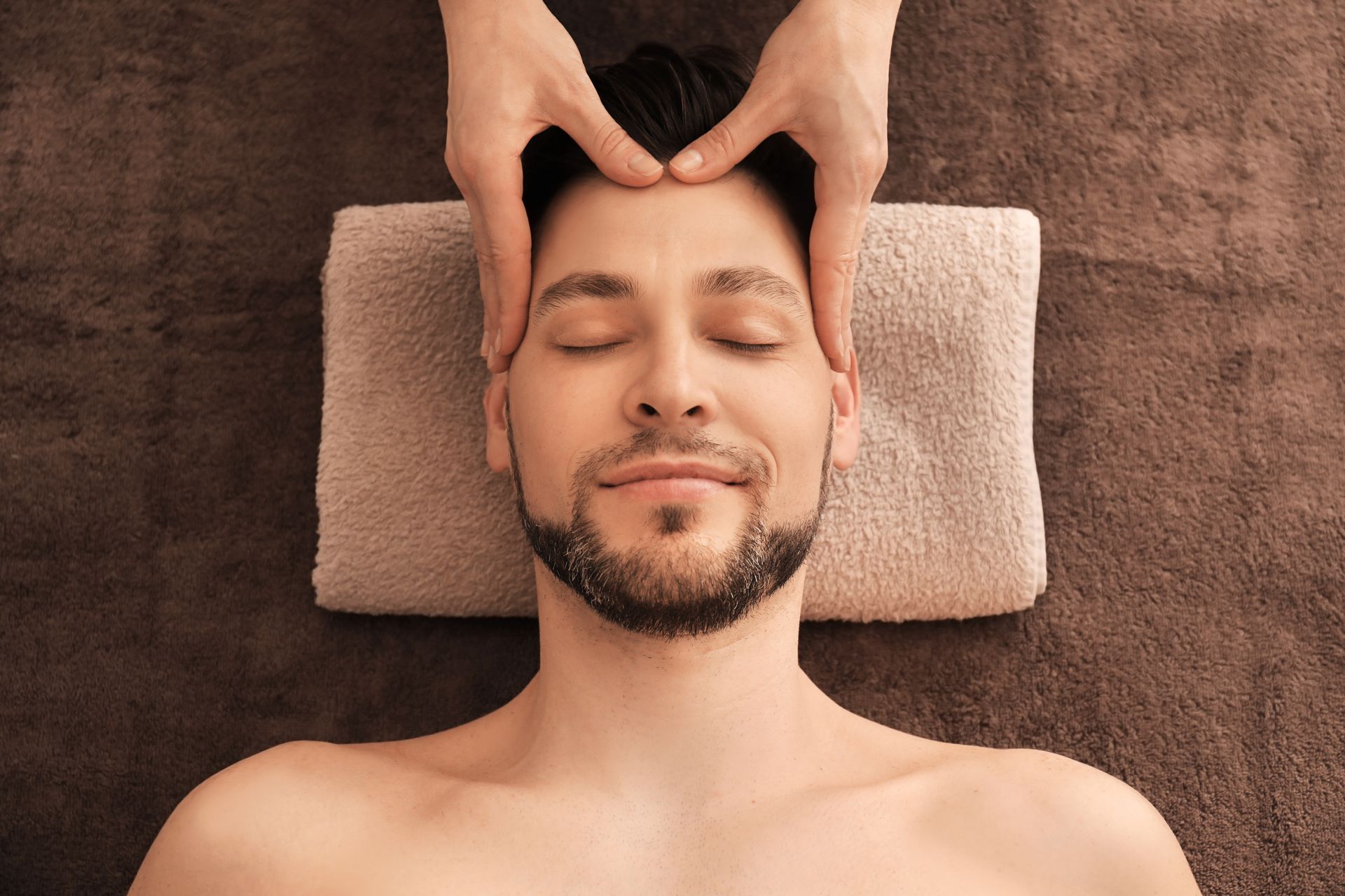 Hands massage a relaxed man’s face on a towel-covered spa table.
