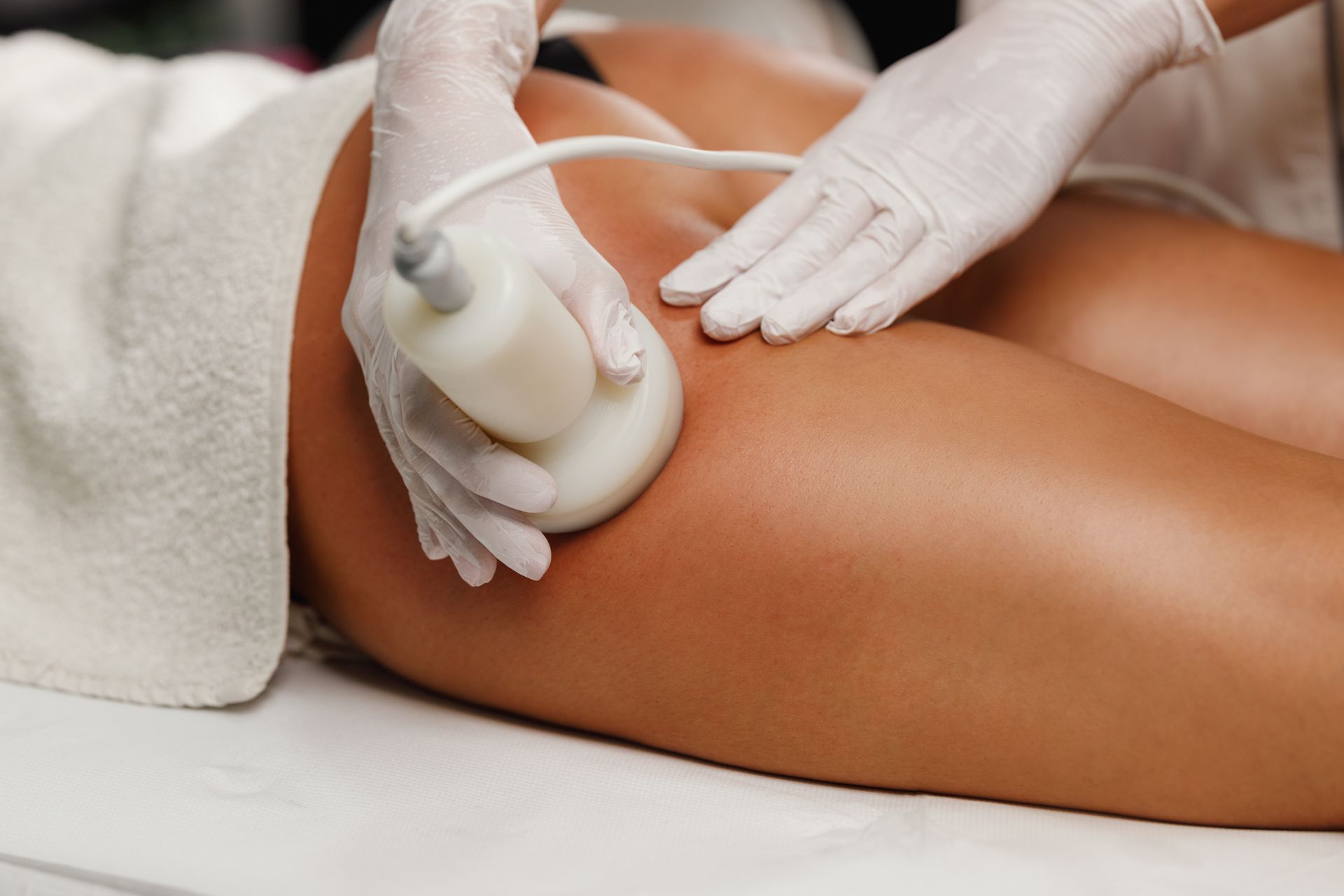 Gloved hands using a white handheld device on a person’s leg during a spa treatment Gloved hands using a white handheld device on a person’s leg during a spa treatment